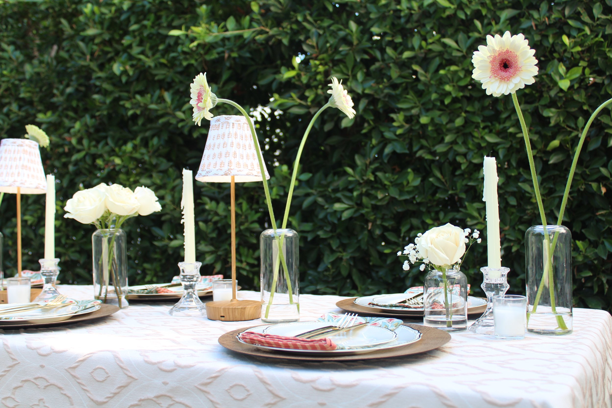 Outdoor table setting with white tablecloth, white flowers in glass vases, candles, and small lamps, against a green leafy background.
