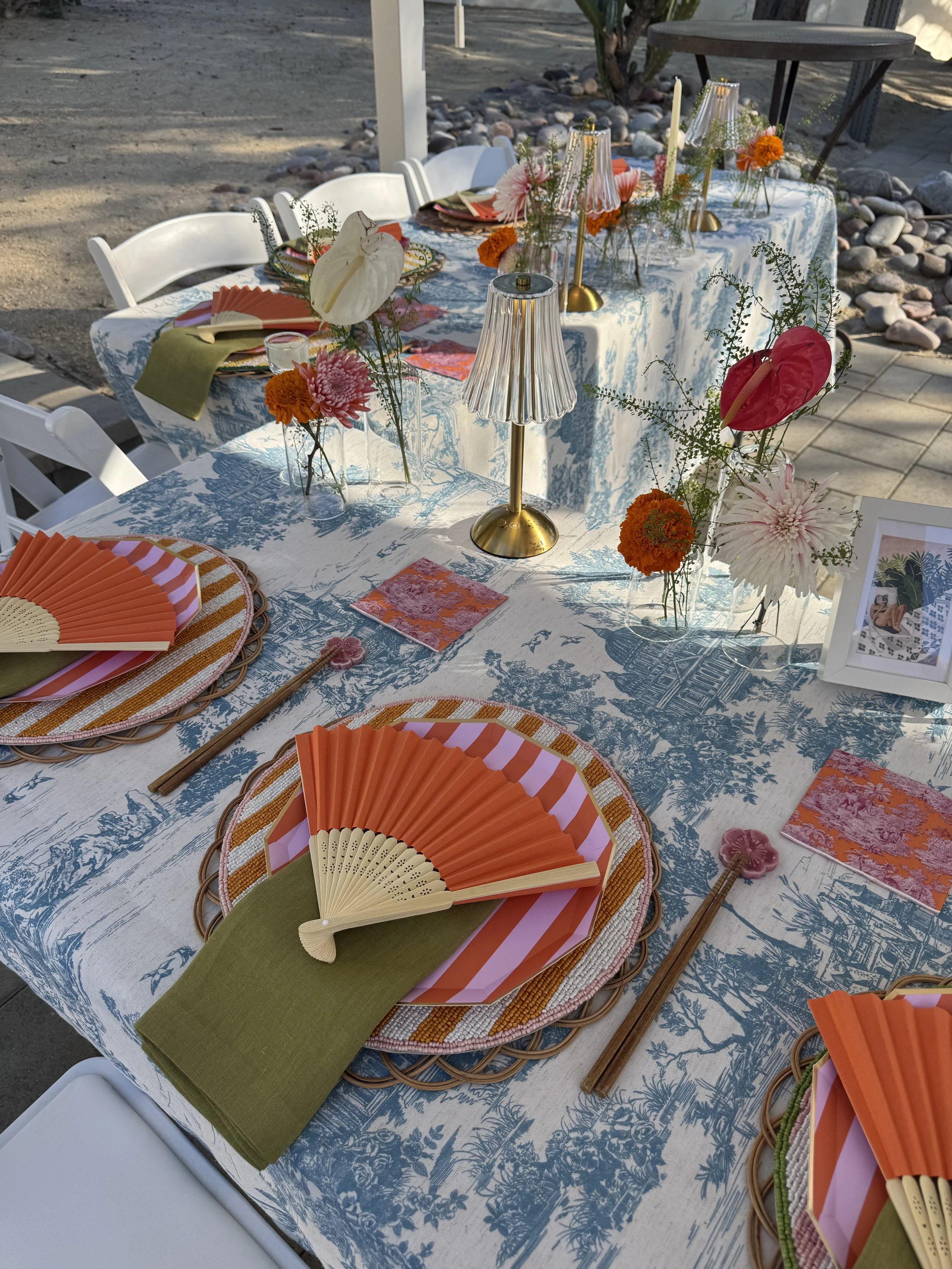 Decorated outdoor dining table with colorful plates, fans, floral centerpieces, lamps, and framed photo, set for a gathering.