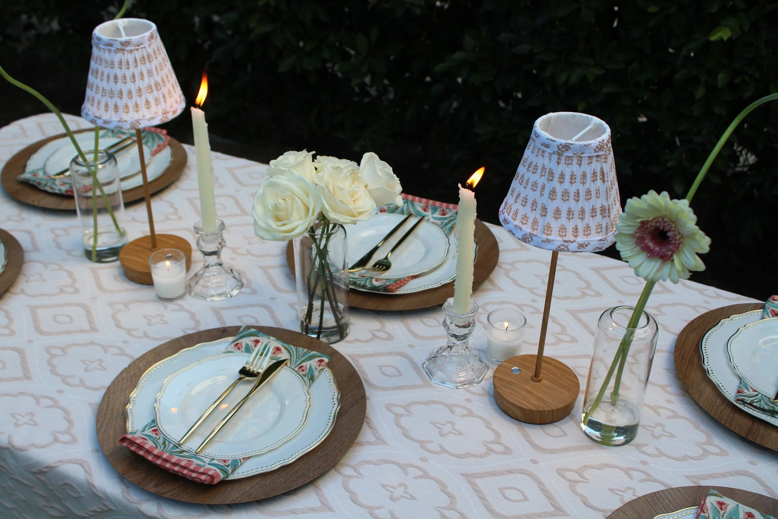 A decorated outdoor dining table with plates, gold cutlery, white flowers, candles, and two small lamps with patterned lampshades.
