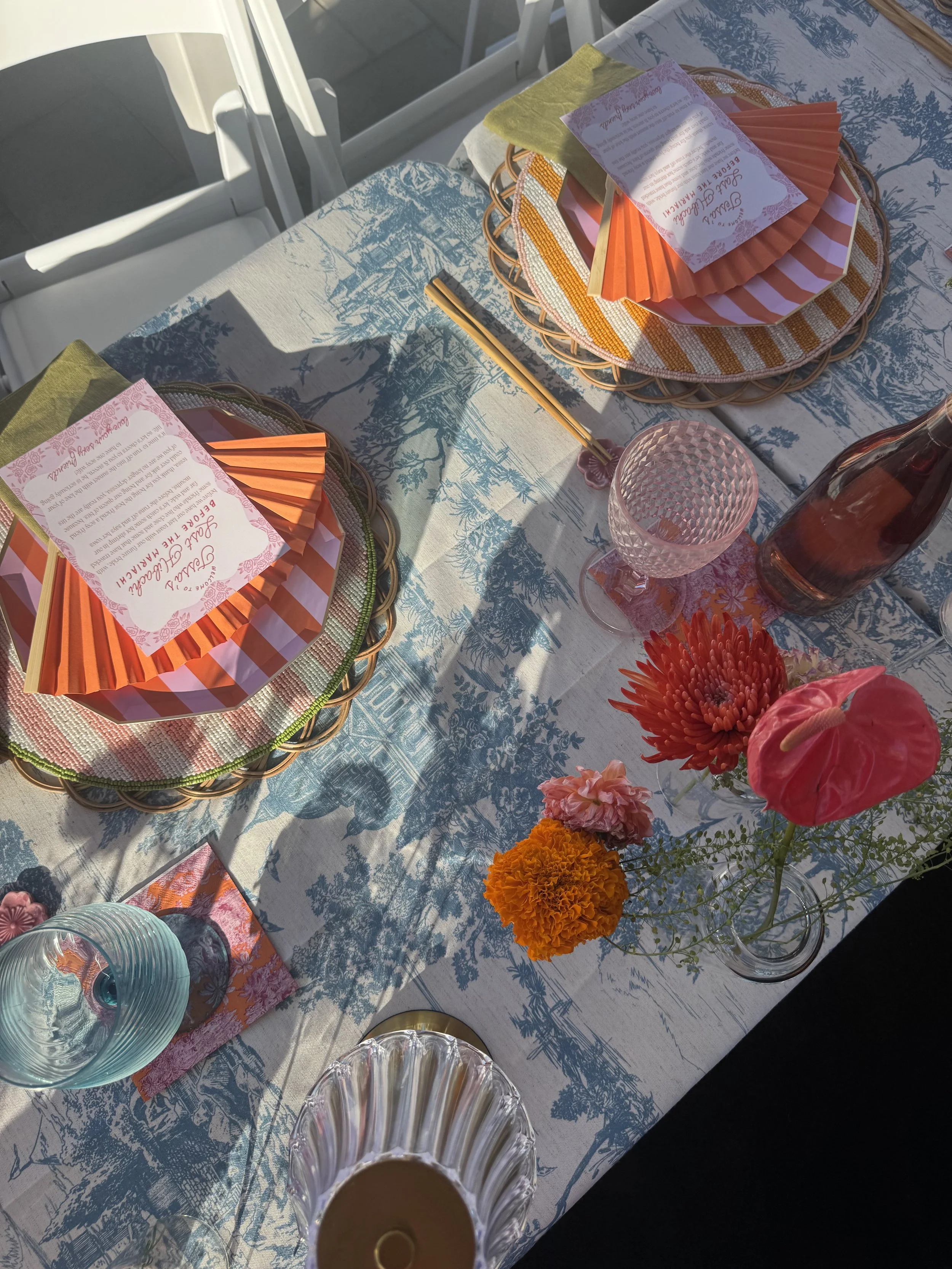 A table setting for a celebration, with floral centerpieces, plates, napkins, and glasses arranged on a tablecloth with a scenic print, illuminated by sunlight.