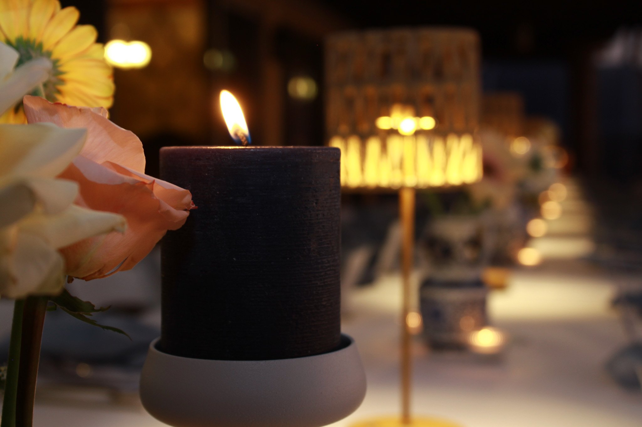 A lit candle on a white candle holder with pink and yellow flowers nearby. The background shows a blurred row of candles and lights.