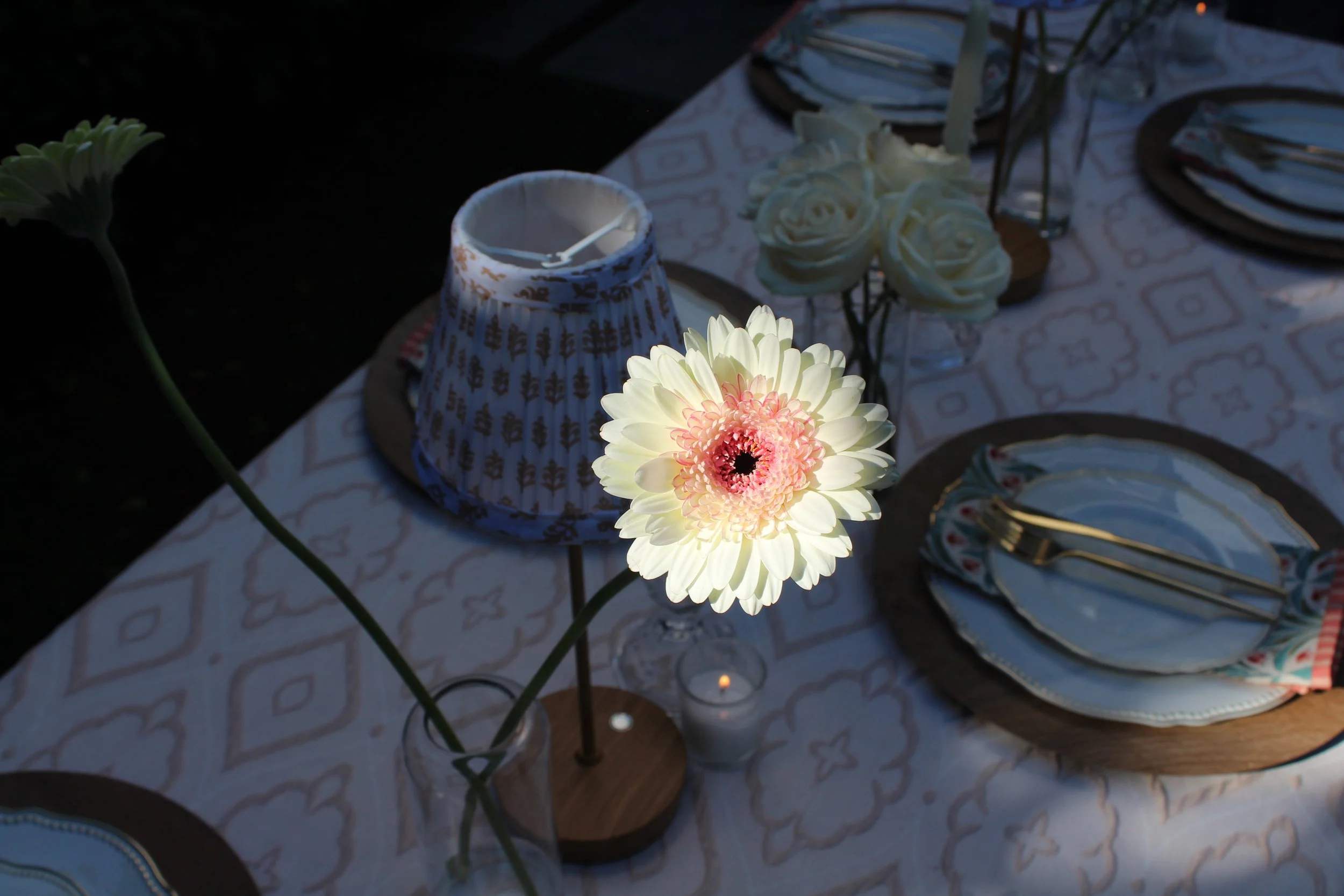 Table set with plates, gold utensils, floral centerpiece, candle, and a small lamp, with a single illuminated flower in the foreground.