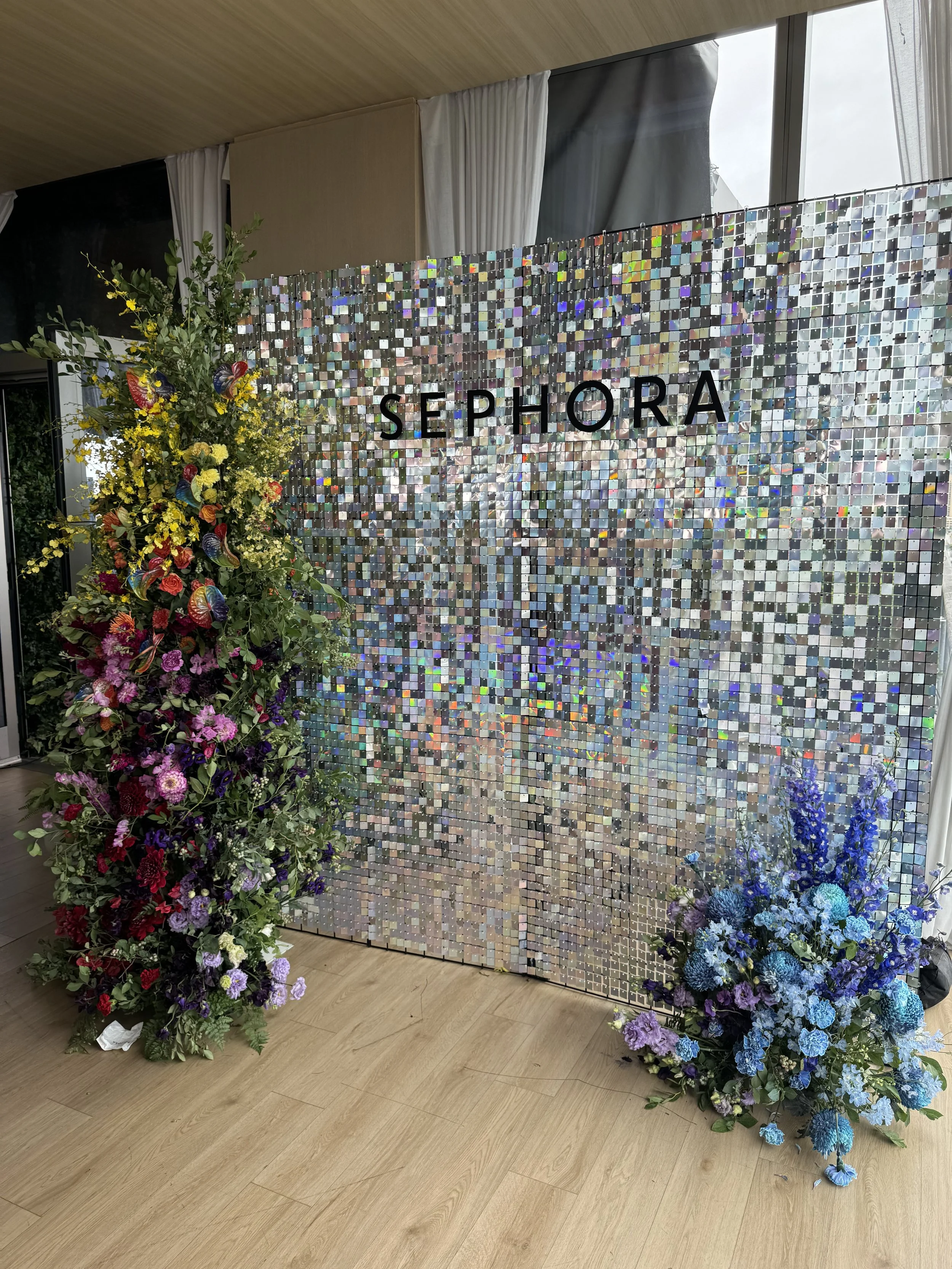 Decorative backdrop with the brand name 'Sephora' and two large floral arrangements, one with yellow, red, pink, purple flowers on the left and the other with blue and purple flowers on the right, in an indoor event space.
