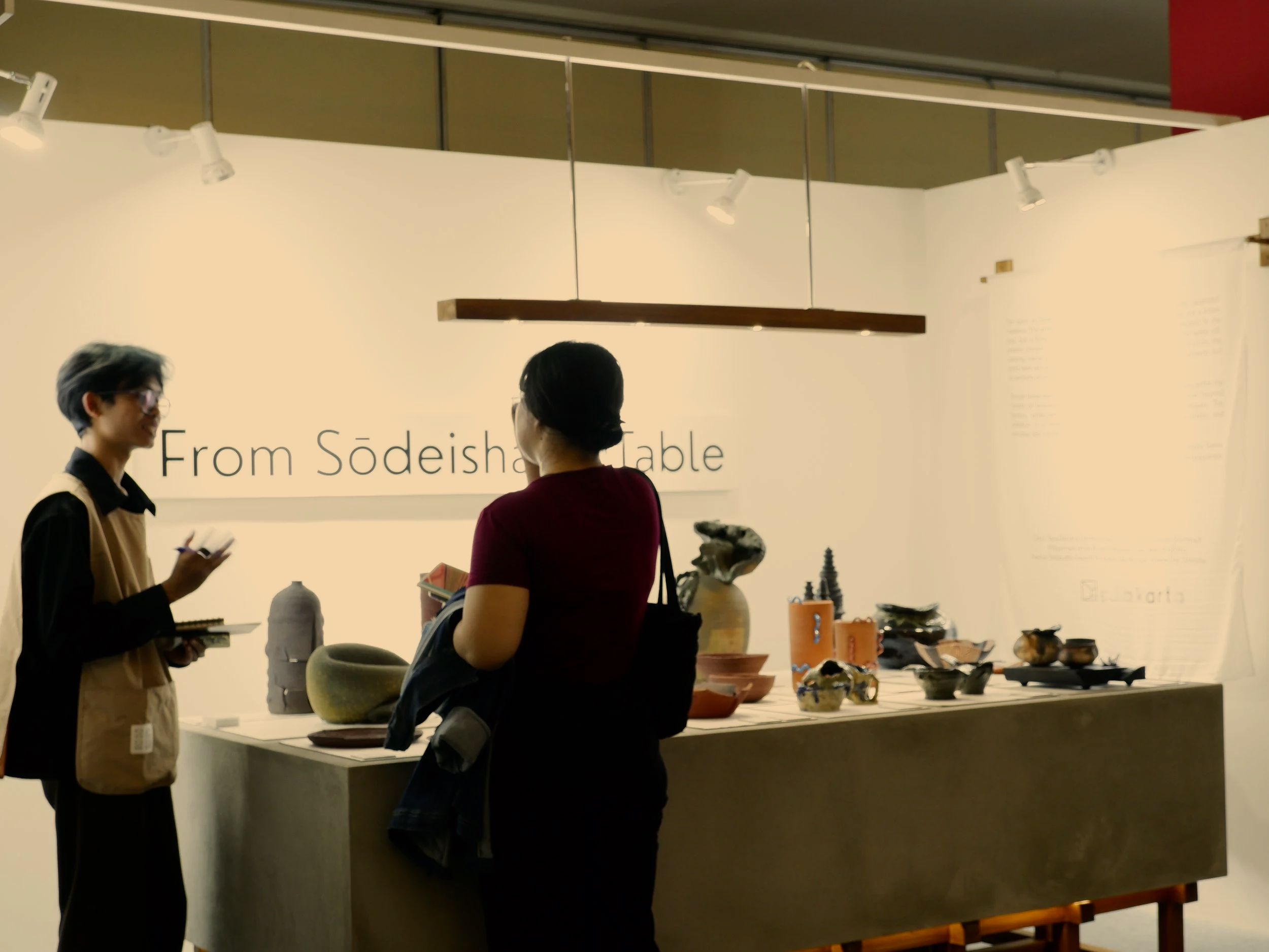 Two women talking at a display of traditional pottery and ceramics in an exhibition, with a sign that reads 'From Södeishanable' in the background.