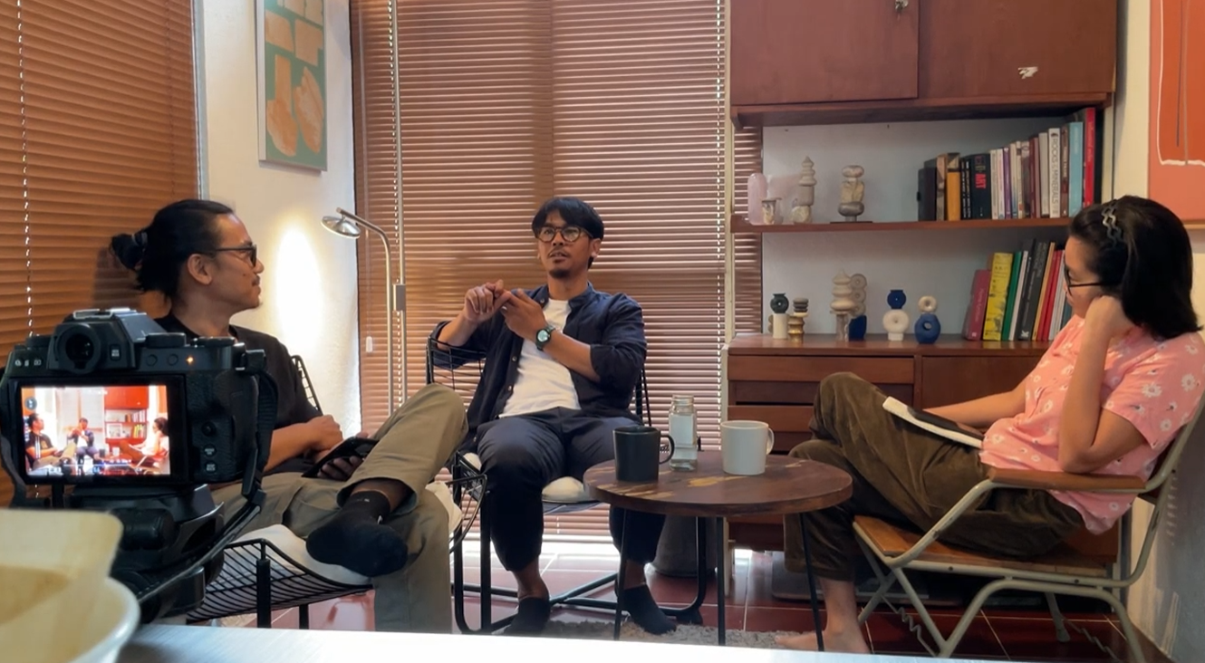 Three people having a discussion in a cozy room with wooden blinds, a bookshelf, and a small round table with cups and a water bottle.