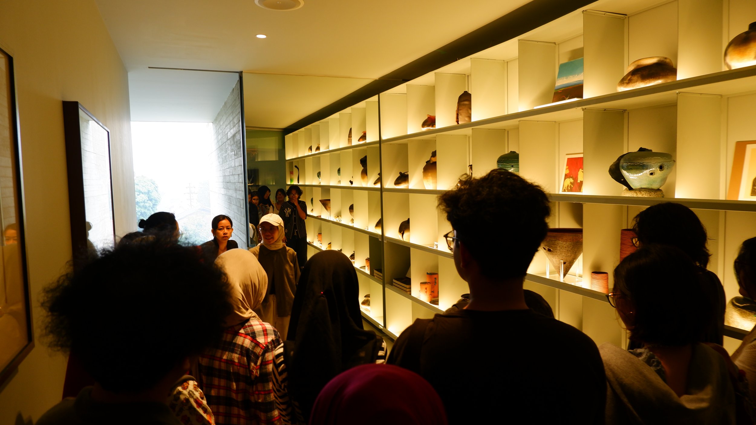 People observing glass art pieces displayed on illuminated shelves in a dimly lit museum.