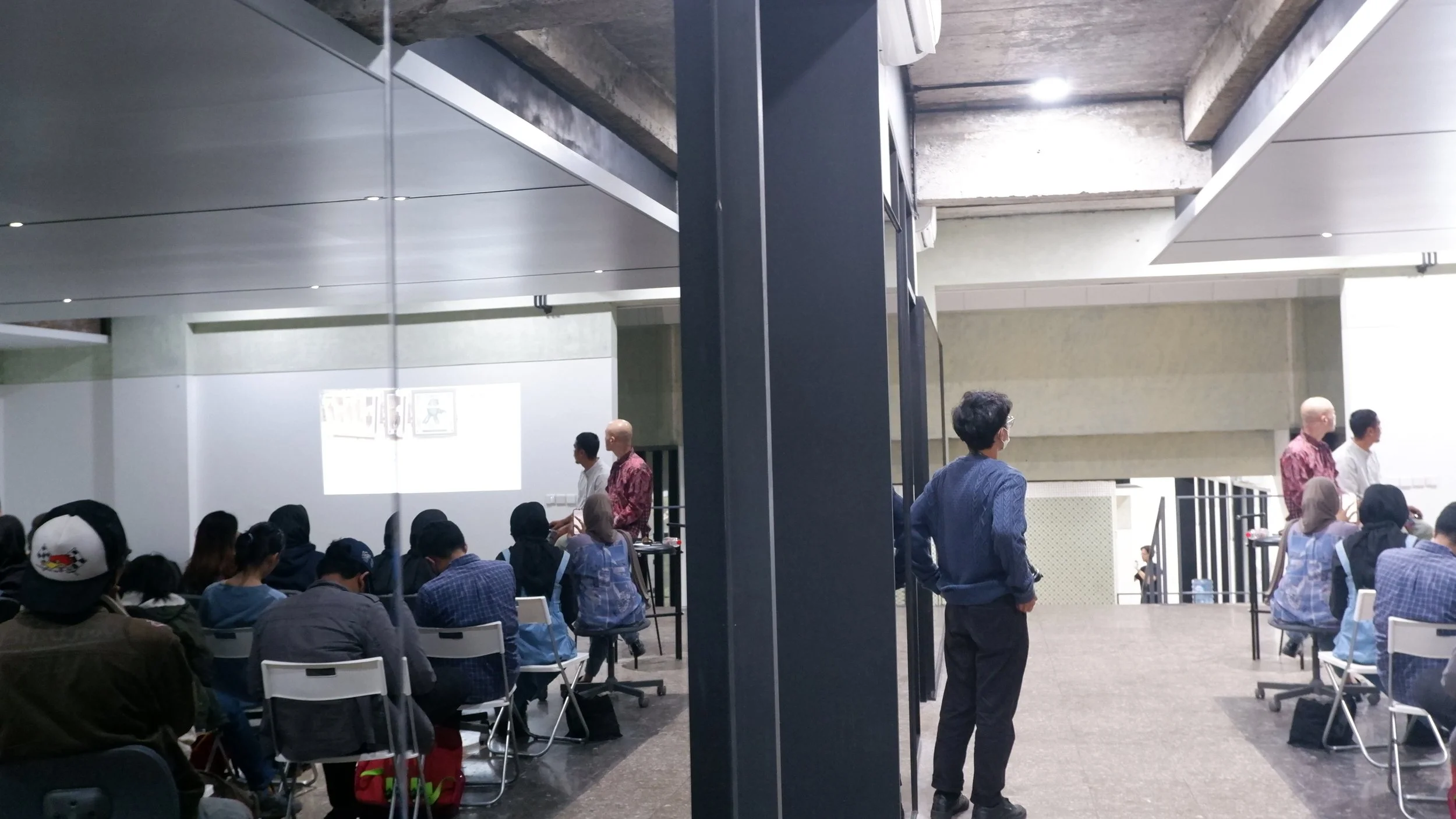 A group of people attending a presentation or workshop in a modern, industrial-style conference room. Several individuals are seated facing a white wall with a projected screen, while three men are standing near the wall, listening or observing.