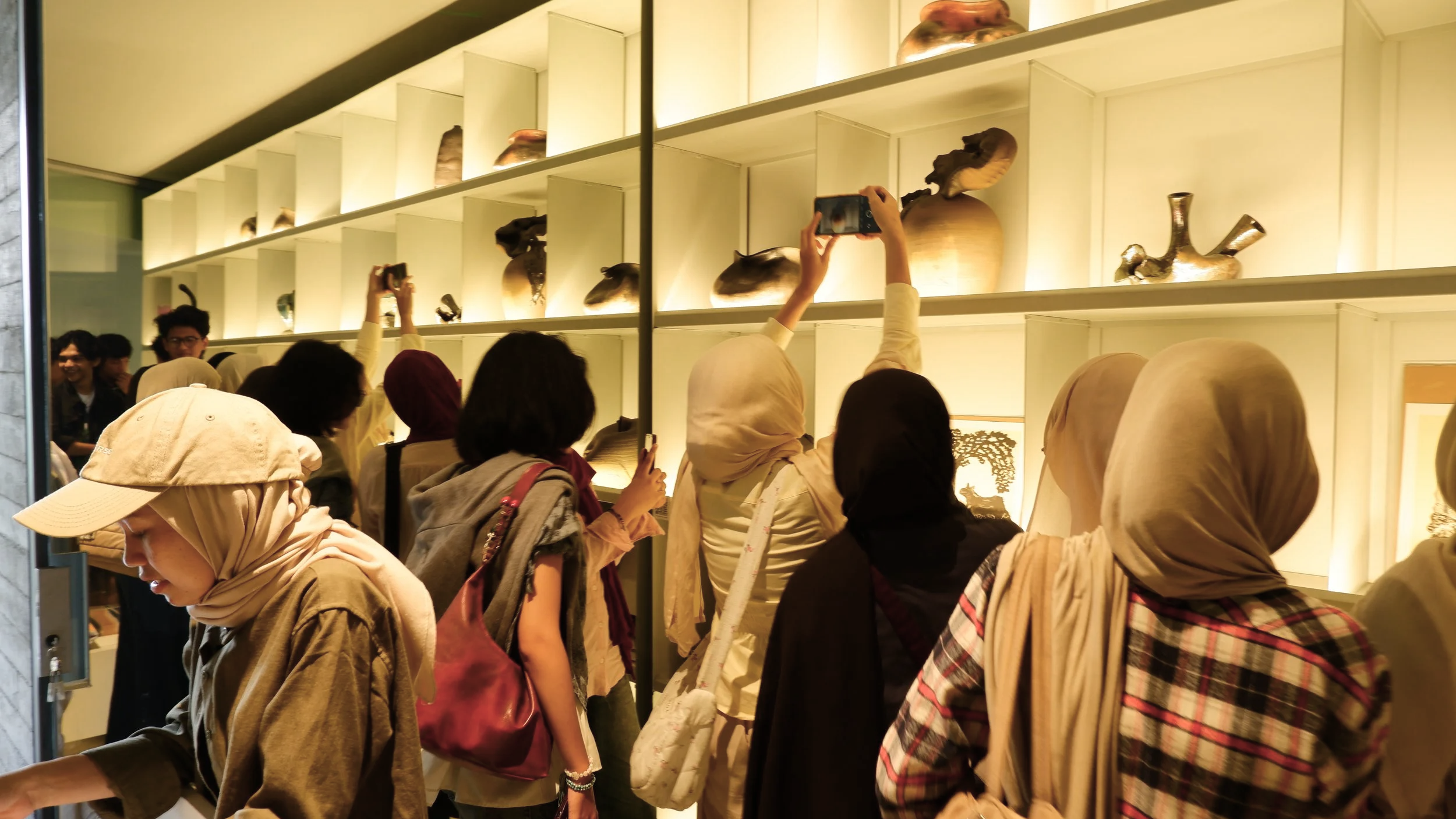 A group of people, mostly women wearing headscarves, are gathered in an art gallery looking at displays of traditional ceramic vessels and sculptures on illuminated shelves.