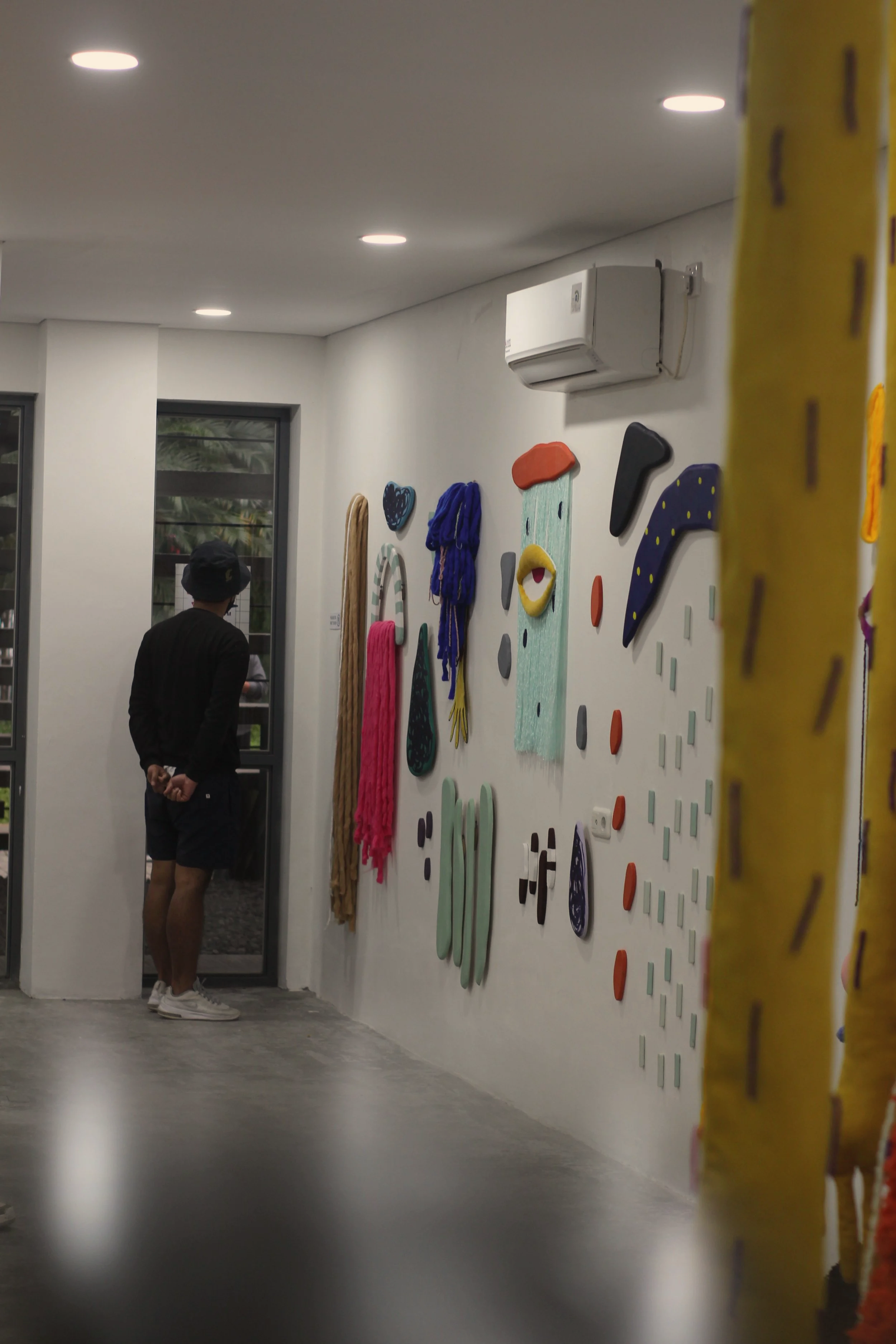 Person wearing a black shirt, shorts, and a bucket hat standing inside near a wall decorated with colorful abstract hanging art pieces and sculptures.