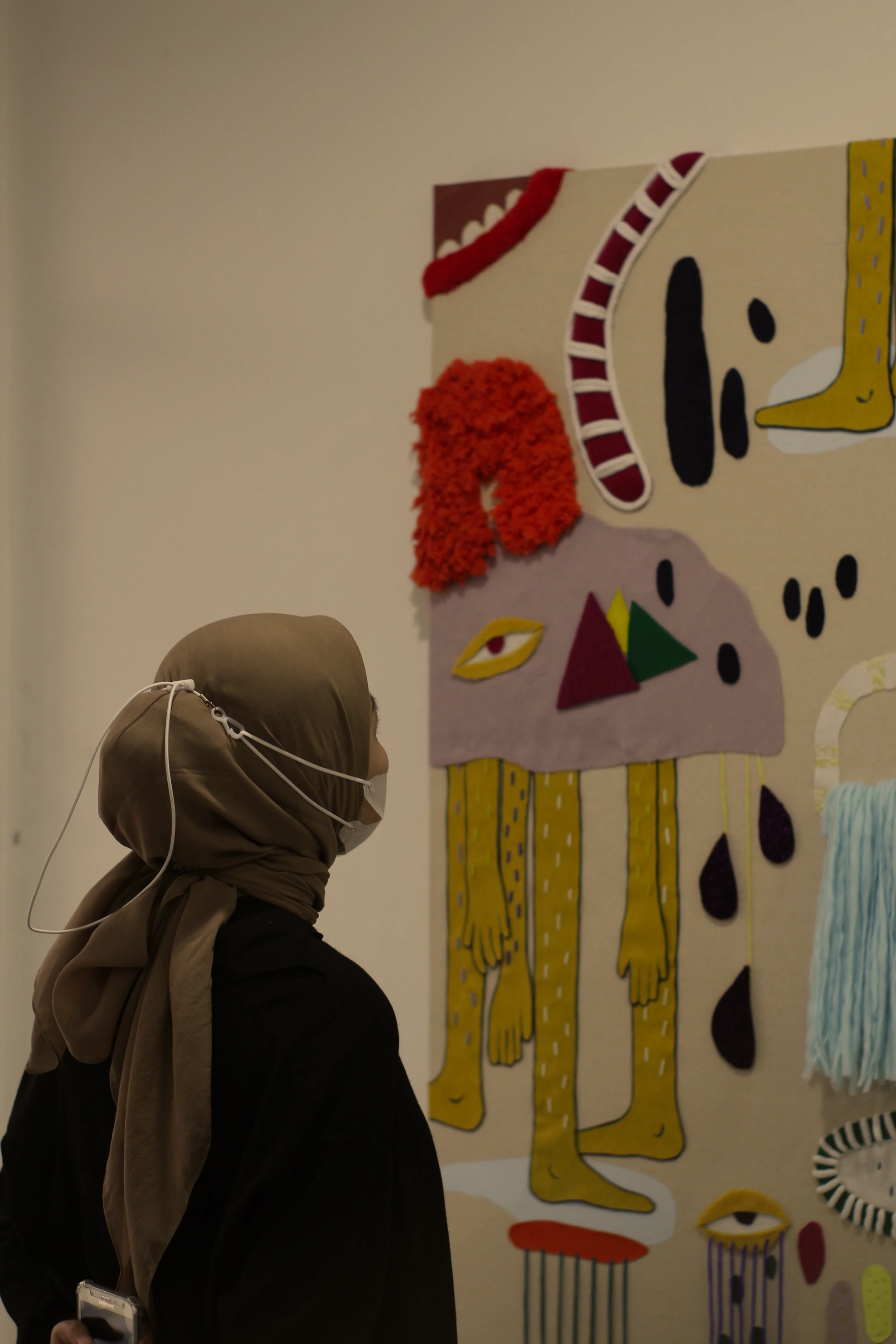 Person wearing a beige headscarf and white face mask looking at colorful abstract artwork.