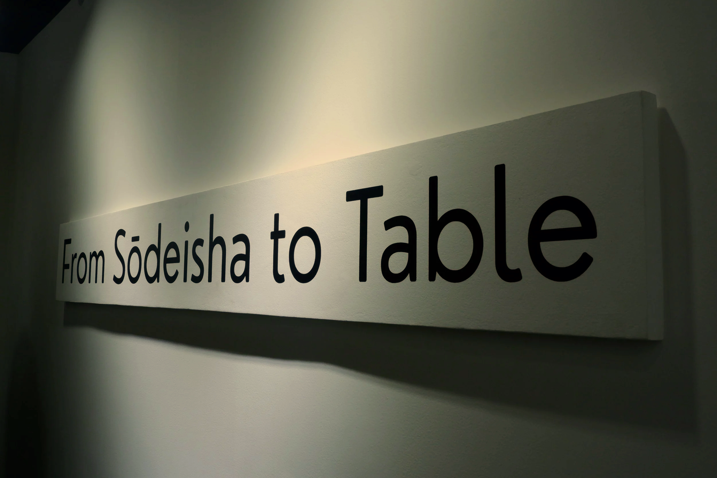 A wall sign that reads 'From Sodeisha to Table' in black text on a white background, illuminated to cast a shadow.