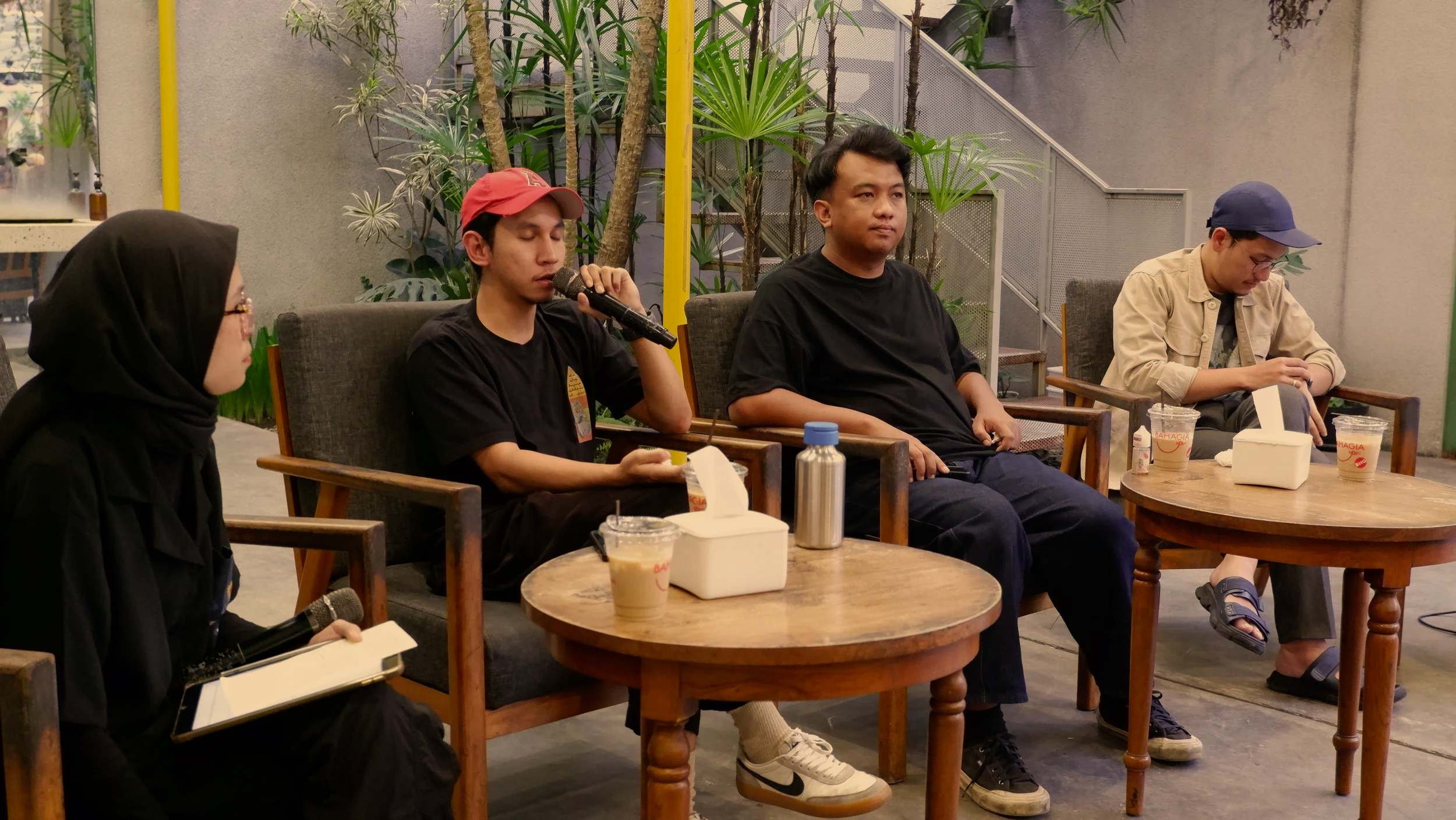 Four people sitting in chairs at a panel or discussion event, with plants behind them and cups on the tables in front of them. The person on the far left is a woman wearing glasses and a black hijab, holding a microphone and a notebook. The second person from the left is a young man with a red cap speaking into a microphone. The third person from the left is a man in a black shirt listening. The person on the far right is a man in a beige jacket and navy blue cap, looking down at his phone.