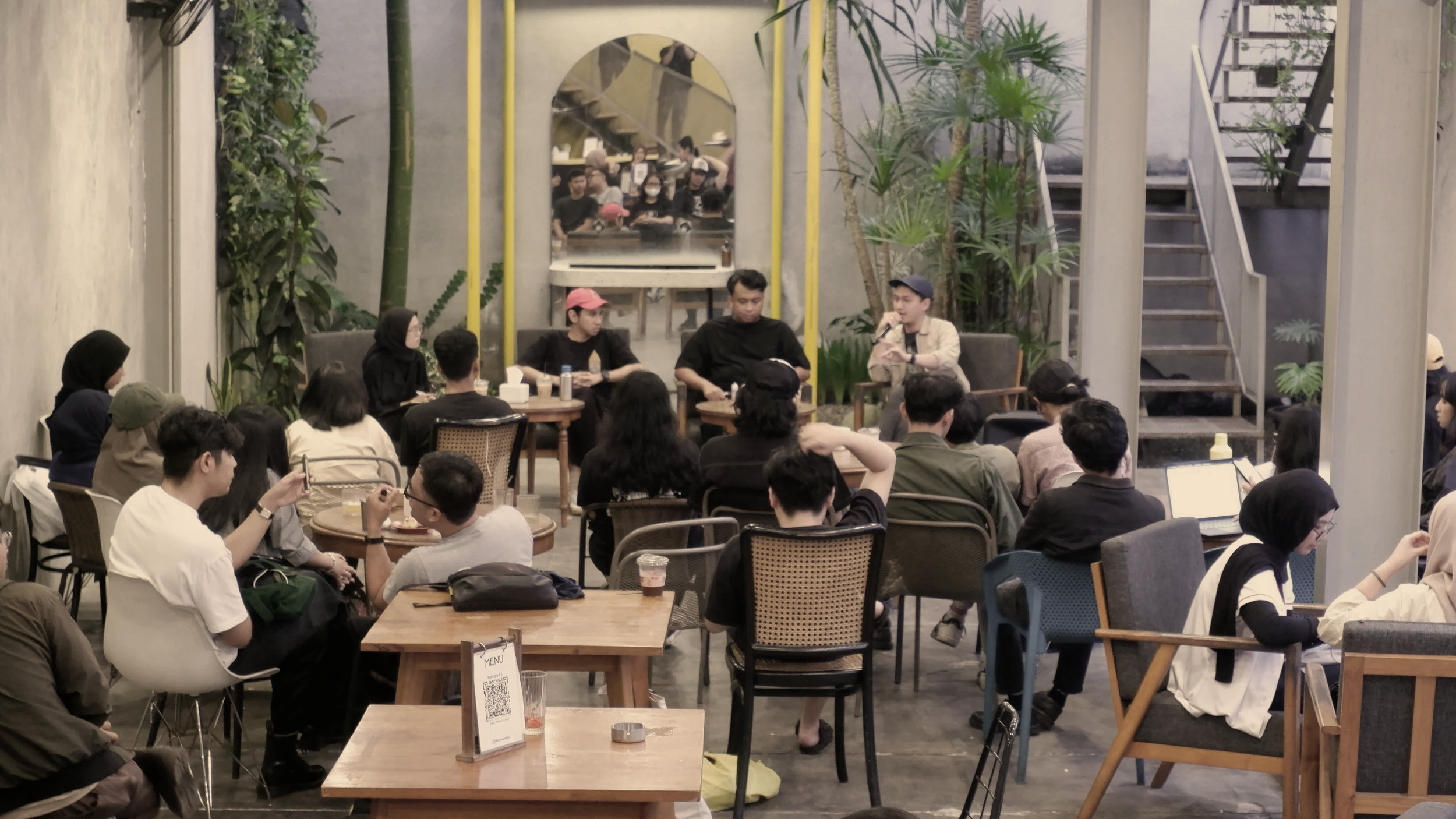 People attending a panel discussion at a cafe or restaurant, sitting at tables, listening to four individuals on a stage. Green plants and decor are visible, as well as a staircase and a mirror reflecting some of the audience.