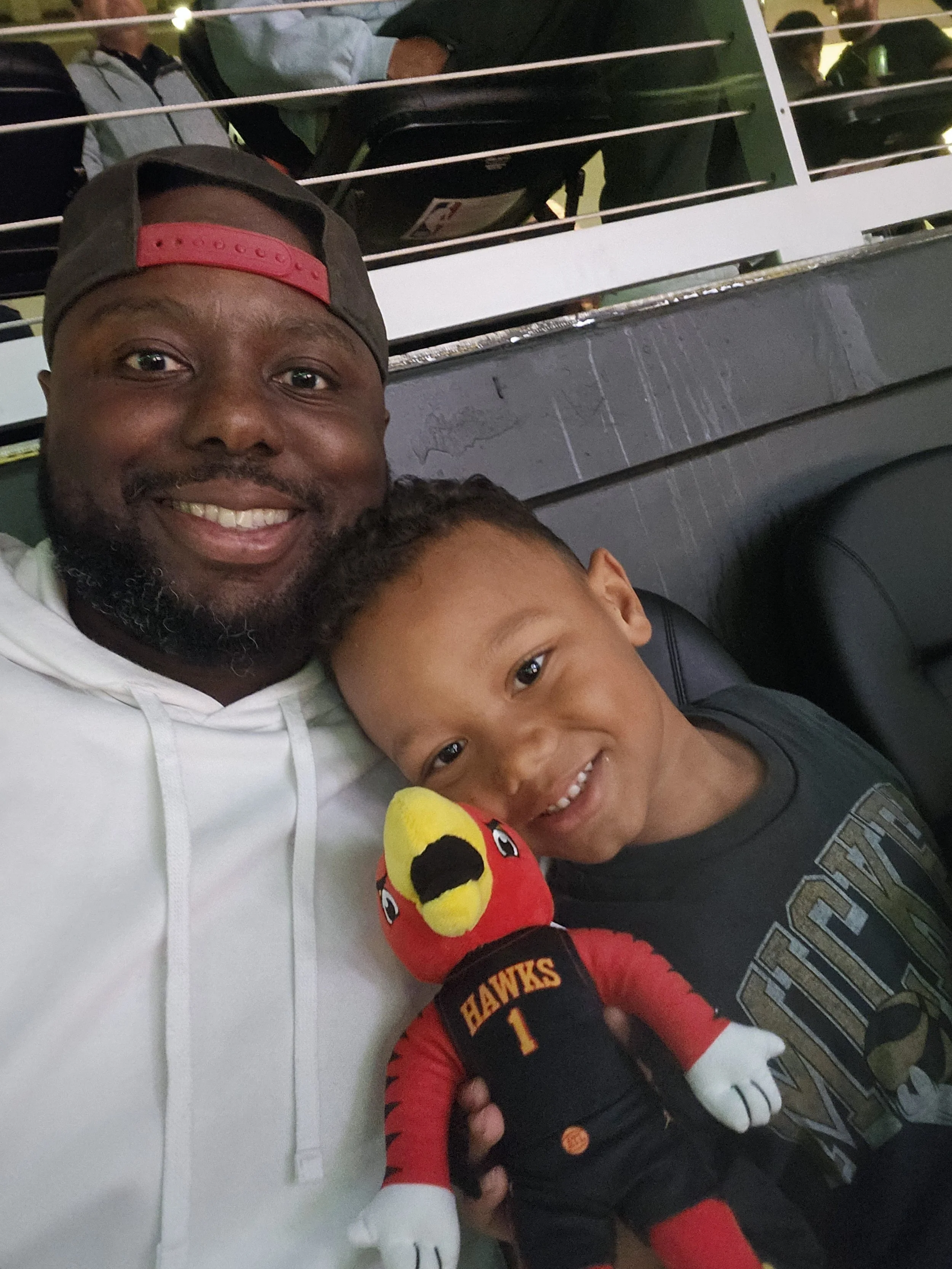 A man and a young boy smiling at the camera, sitting together at a sports stadium. The boy is holding a plush toy of a red bird wearing a black sports jersey with 'HAWKS' and the number 1 on it.