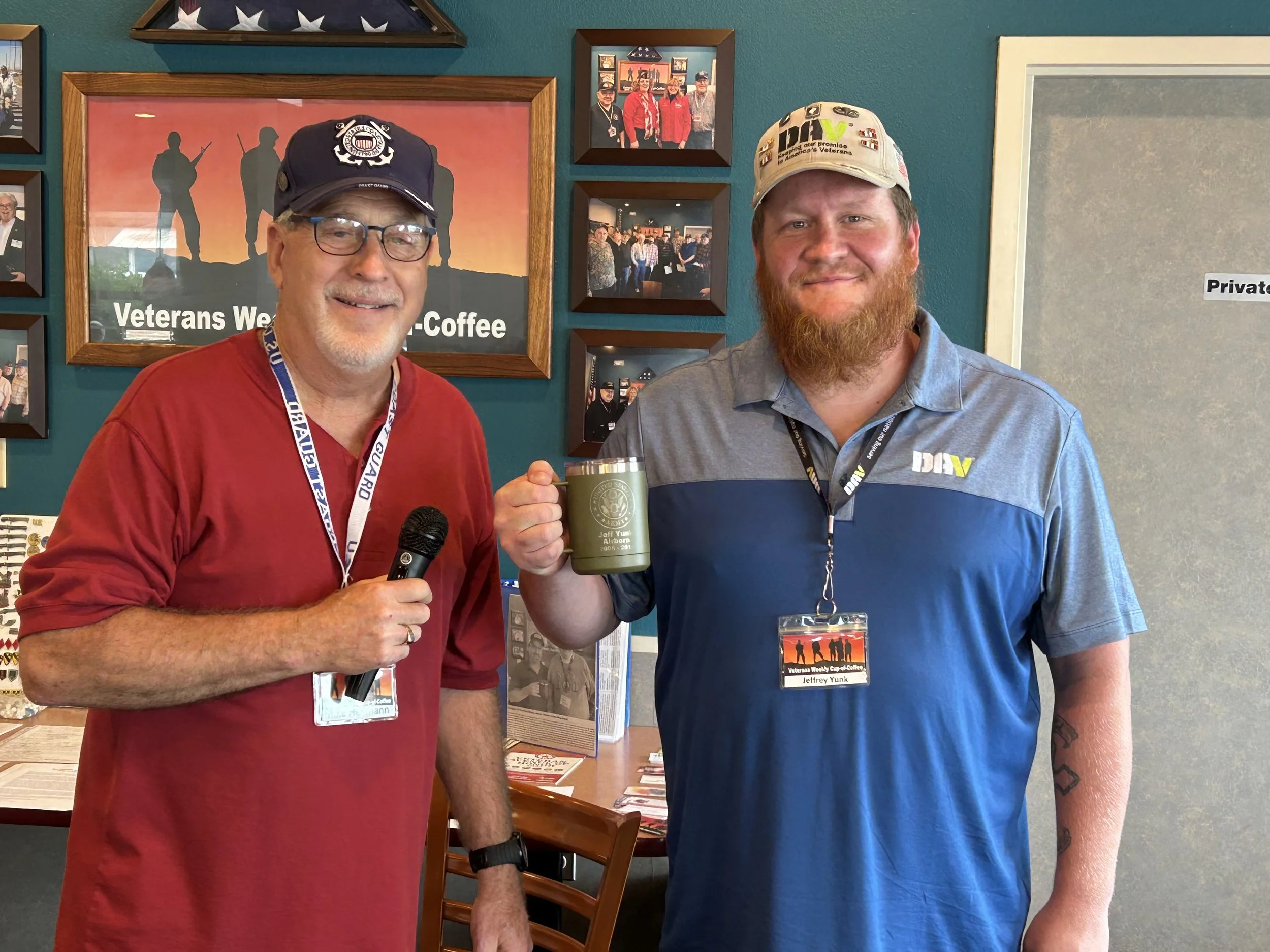 Two men smiling, one holding a microphone and the other holding a green coffee mug, standing in front of a wall with framed photos and a sign that says 'Veterans Weekly Coffee'.