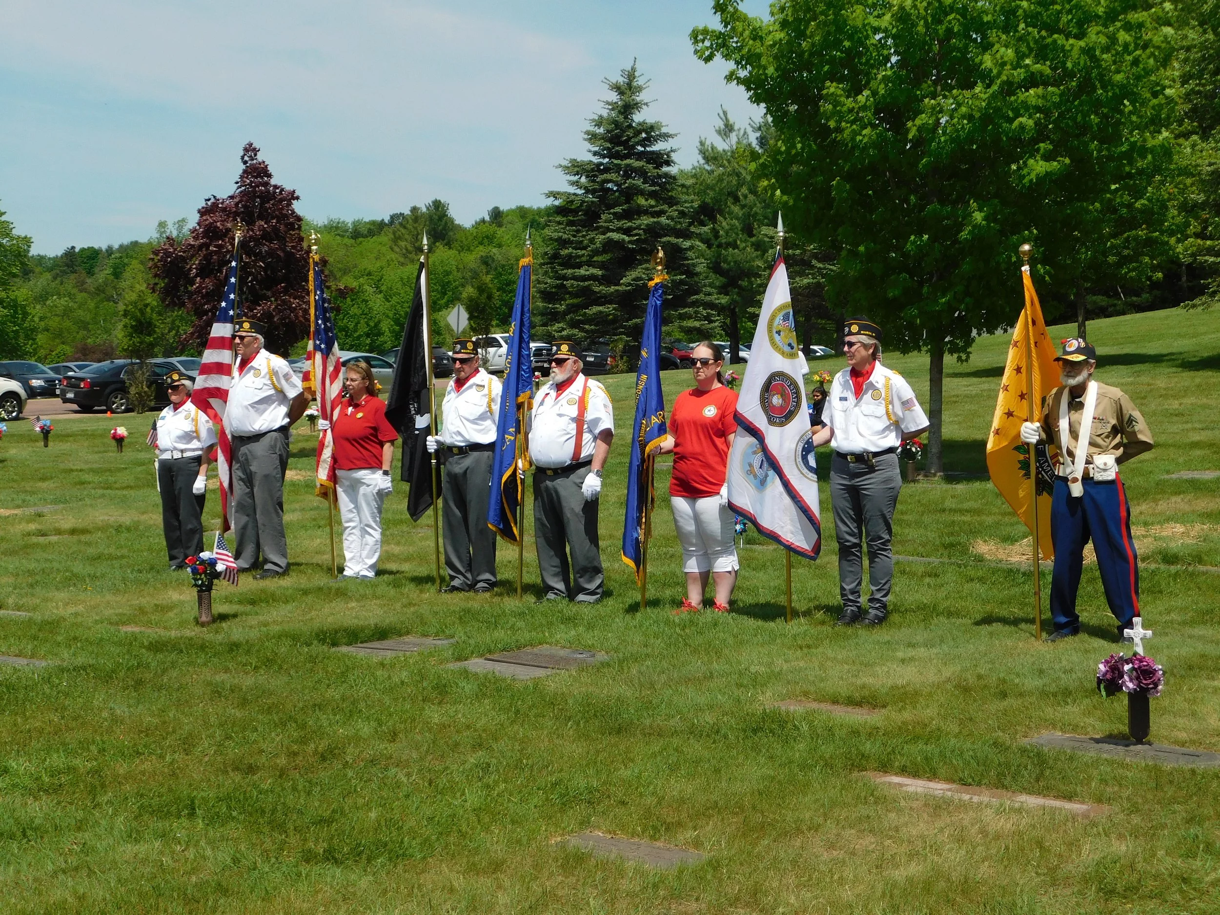 Group of people standing in a line holding various flags at a cemetery during Memorial Day. They are wearing white shirts, red shirts, and military uniforms. Some have medals and hats, with memorial markers and flowers visible on the grass.