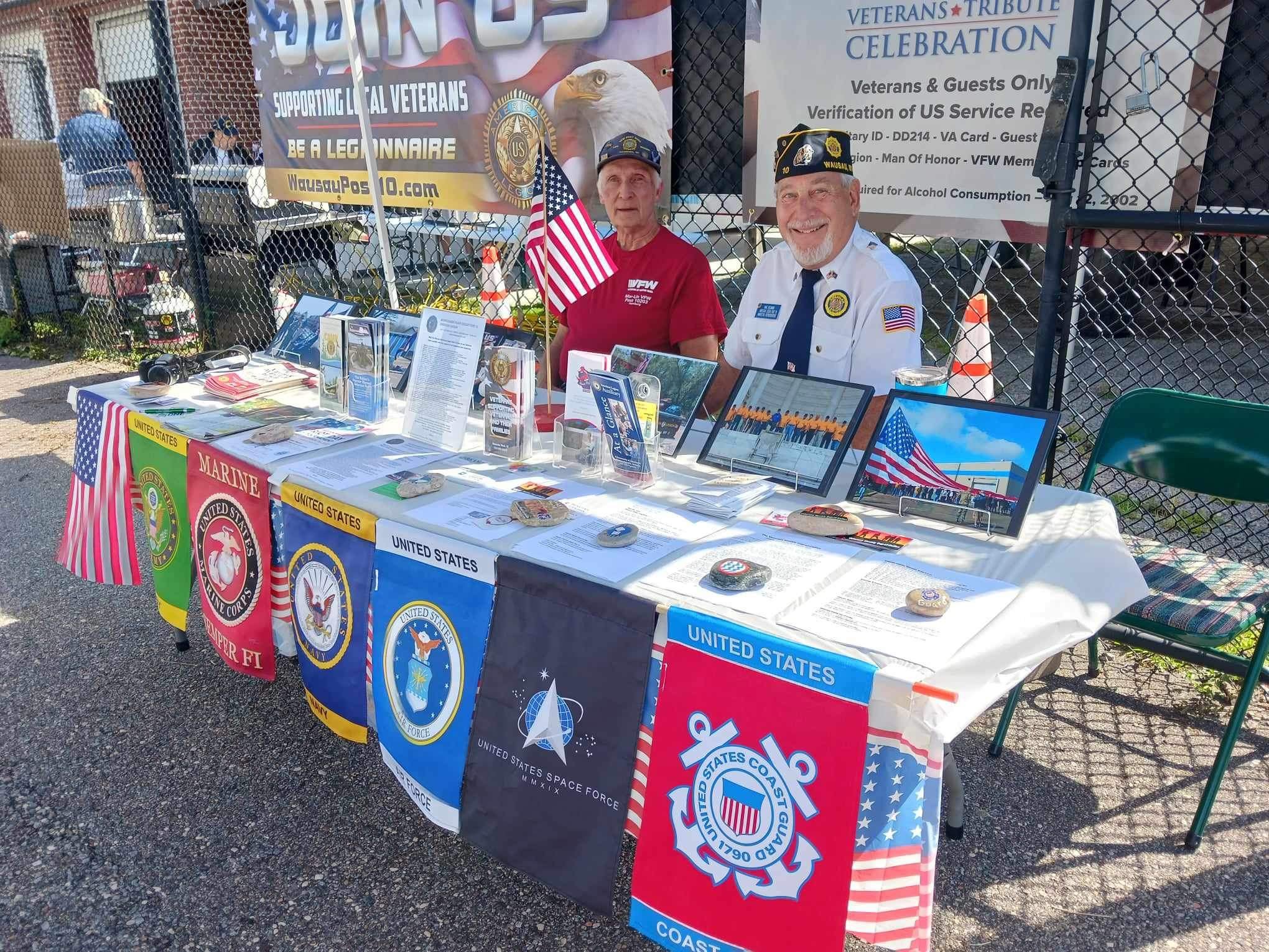 Two men dressed in veteran uniforms are sitting behind a table decorated with various military flags, memorabilia, and informational displays. The table is set up outdoors, enclosed by a chain-link fence, with a large banner in the background indicat