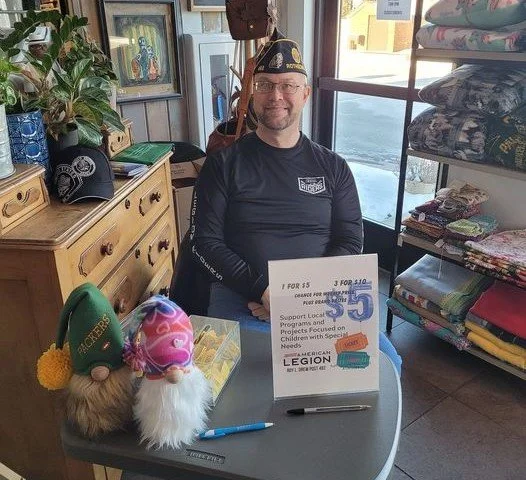 Man smiling, seated at a table with a sign promoting a fundraiser, inside a retail store filled with clothes and decorative items.
