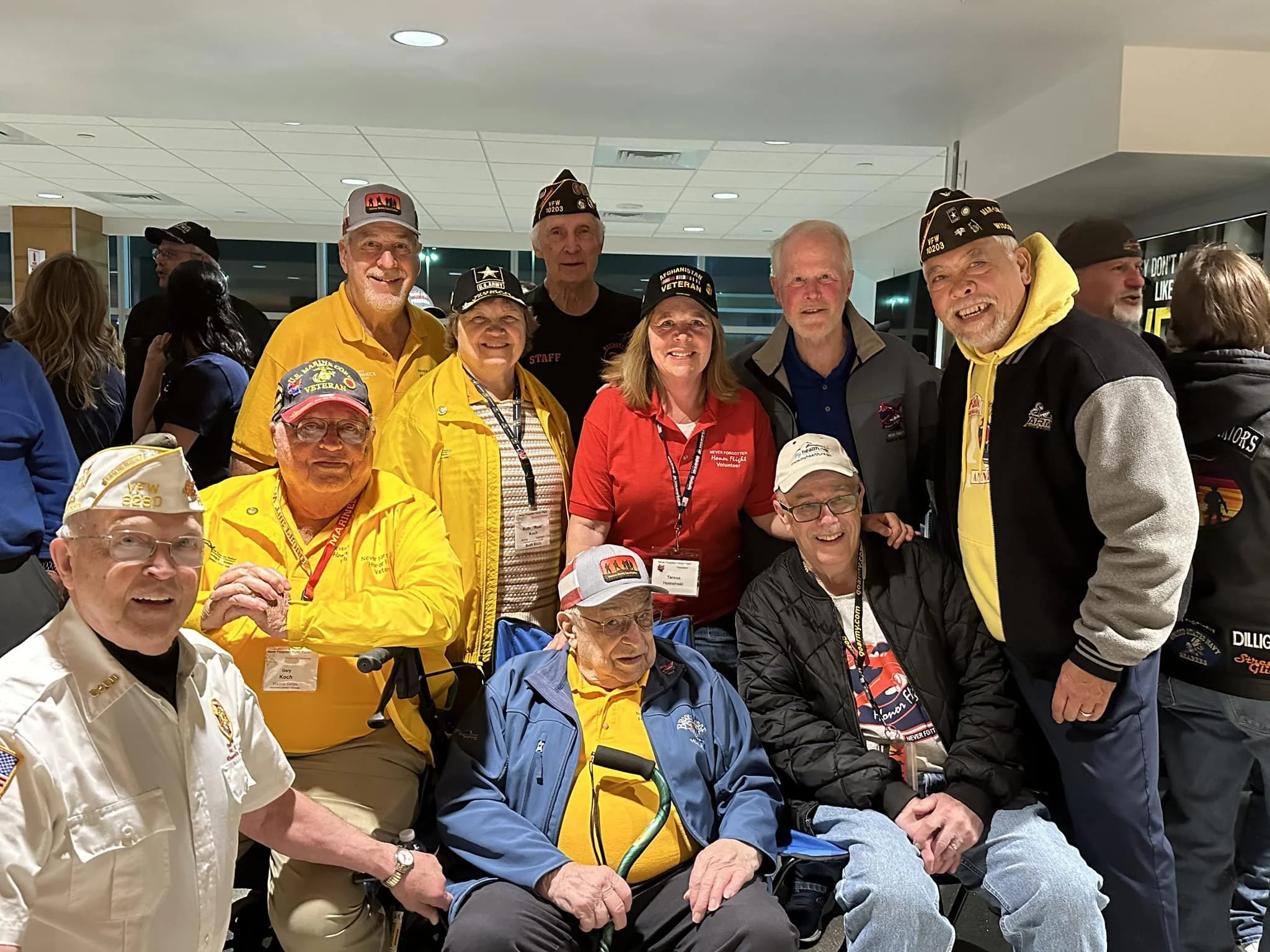 Group of eleven people, mainly veterans, gathered indoors at a reunion or event, some wearing veterans' hats and lanyards, smiling for the camera.