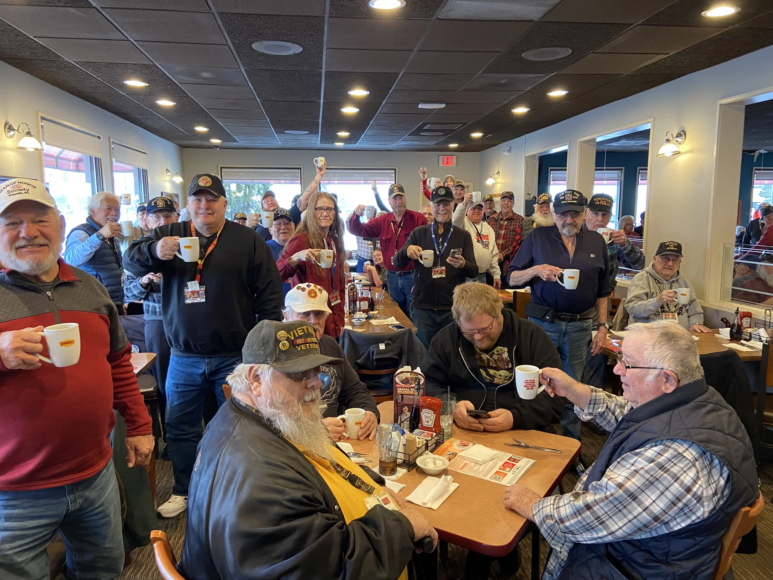 Group of veterans and friends gathered in a restaurant celebrating, holding coffee mugs and smiling.