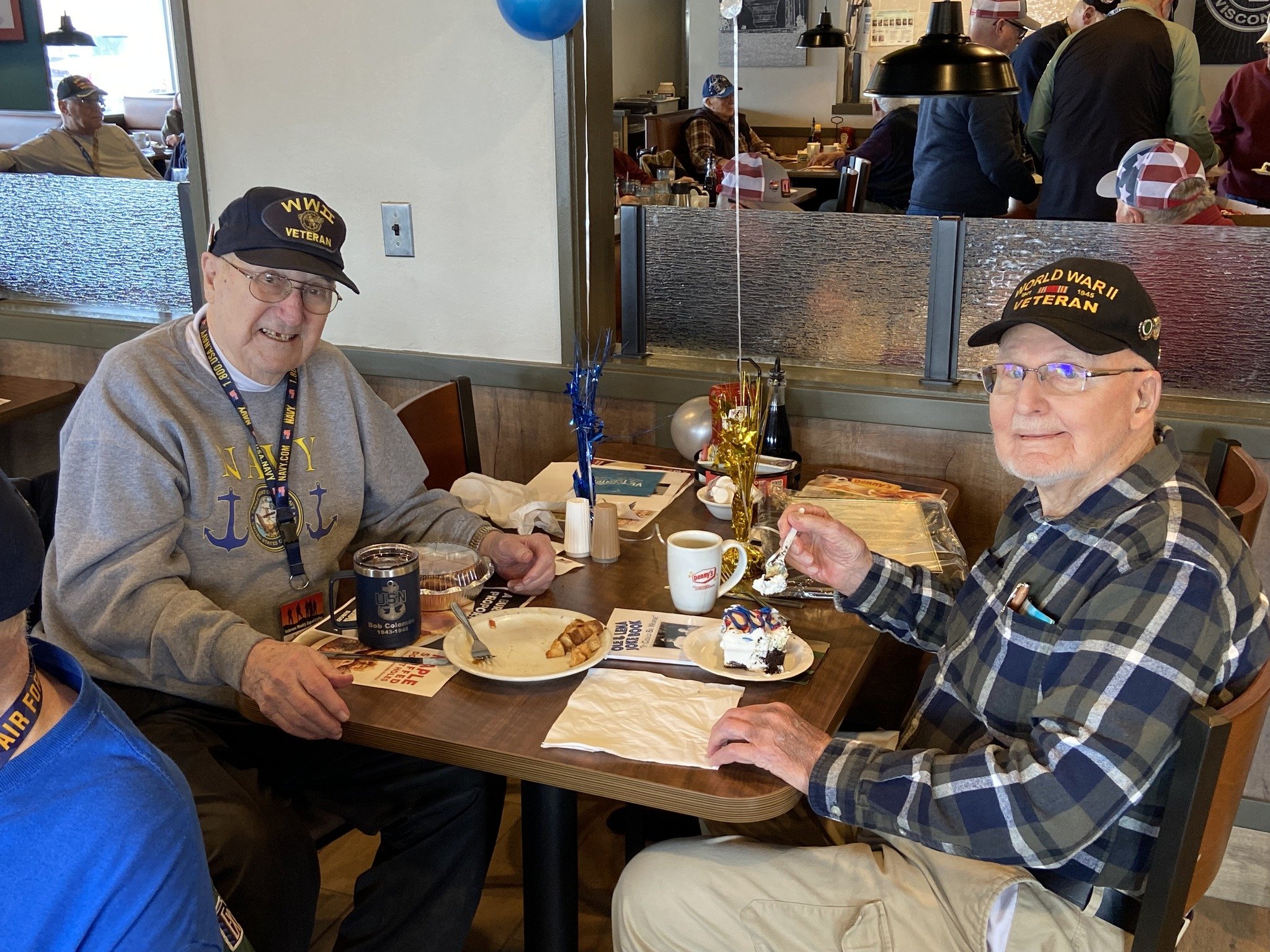Two elderly men in veteran hats sitting at a restaurant table with desserts and drinks, celebrating a special occasion.