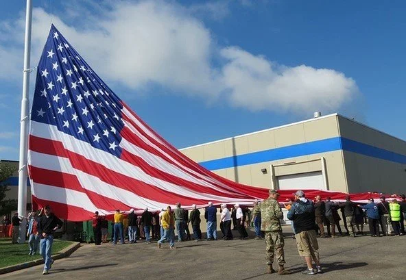 People holding and displaying a large American flag outdoors near a building, with a clear blue sky overhead.