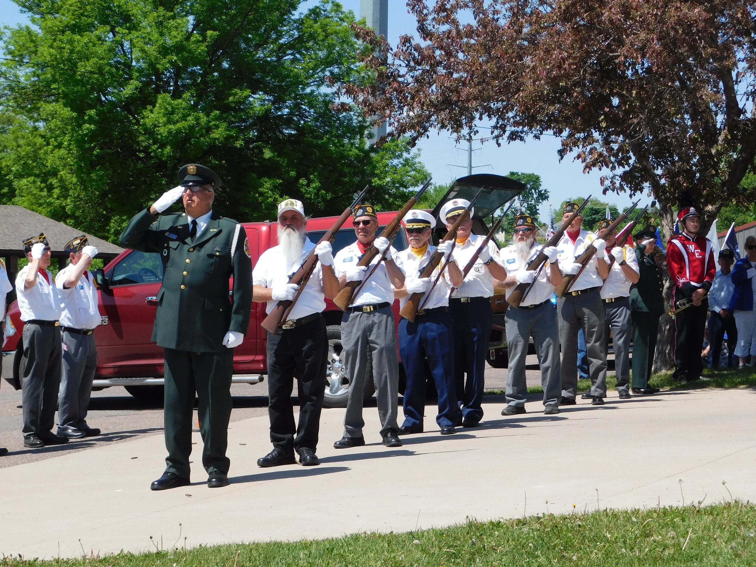 Veterans parade with a man in a military uniform saluting and others holding rifles, some wearing white shirts and hats, under trees on a sidewalk.