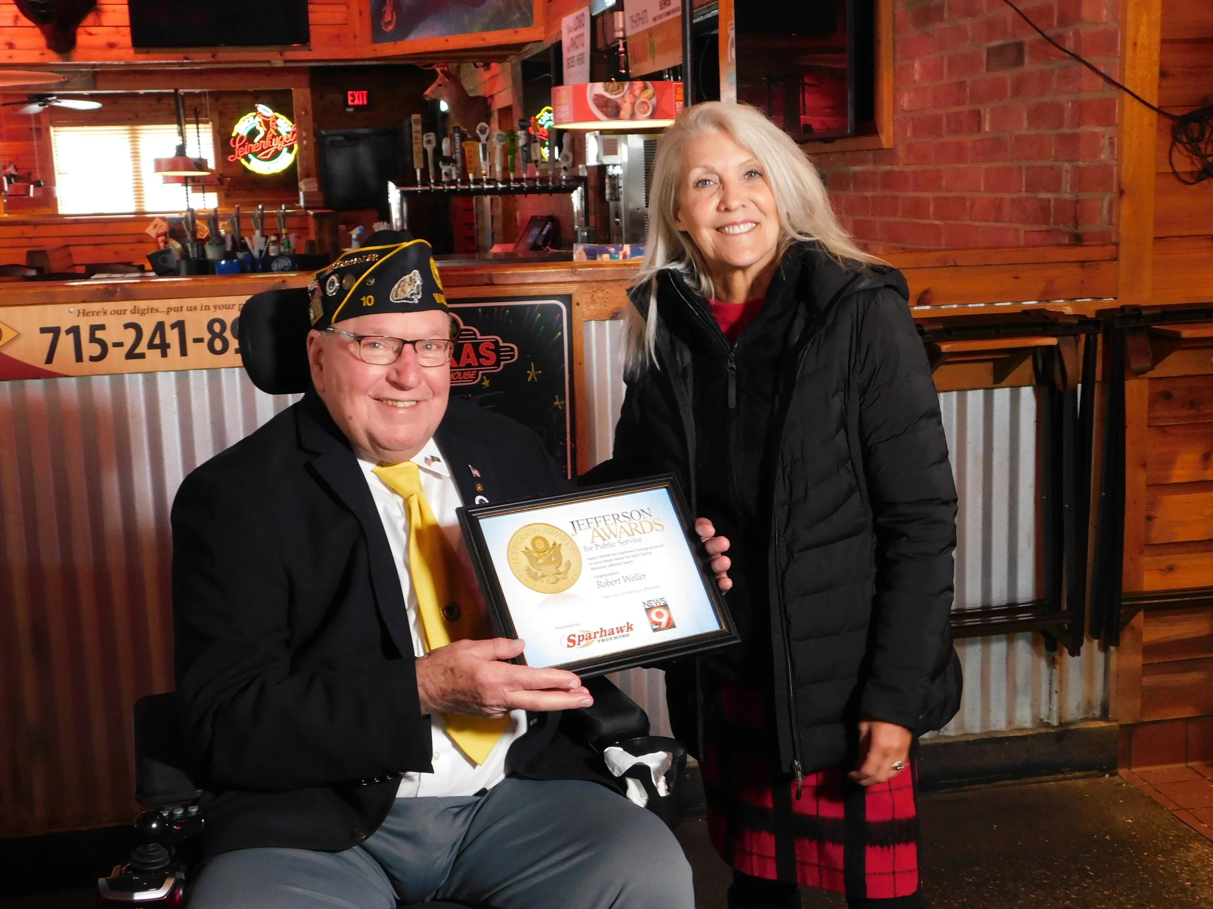 A man in a wheelchair holding a framed award, smiling, wearing a black band with a badge and a yellow tie, standing next to a woman with long gray hair, smiling, wearing a black jacket and red plaid skirt, inside a restaurant or bar with brick walls 