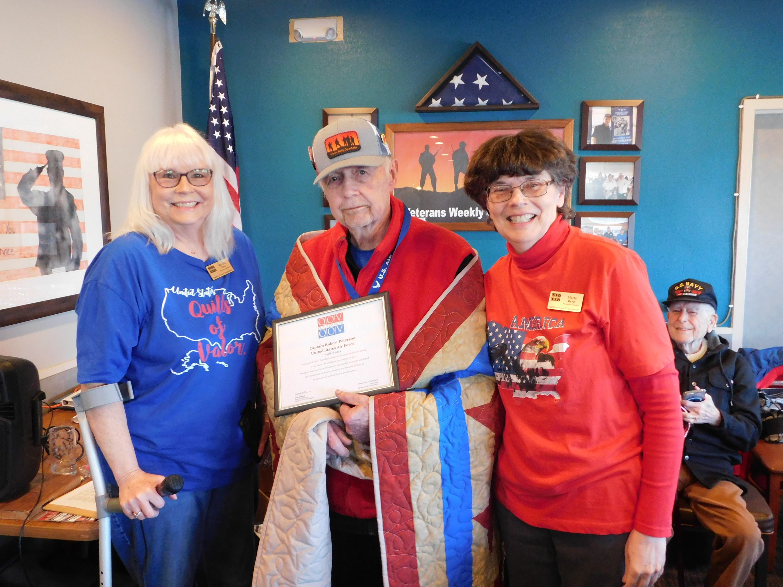 Three women and one man indoors at a veteran recognition event, with patriotic decorations including an American flag and framed photos on the wall. The man is holding a framed certificate, and the women are wearing shirts with patriotic themes.