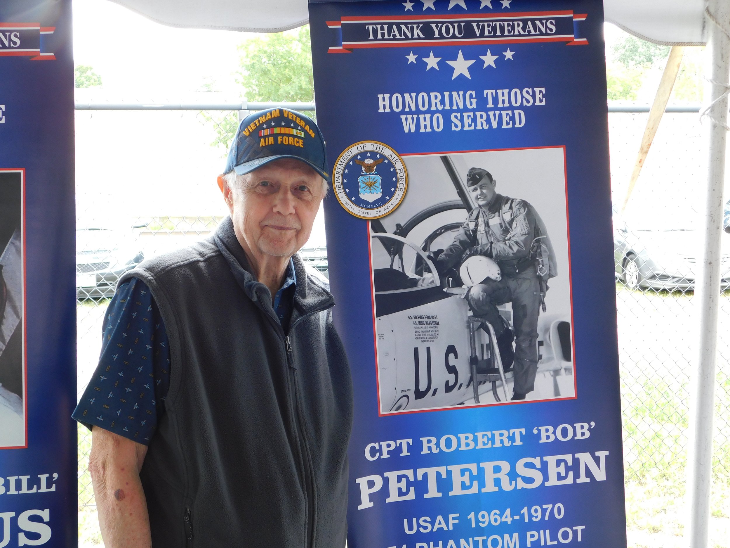 An elderly man wearing a cap and vest stands in front of a blue veterans' appreciation banner at a memorial event. The banner honors Captain Robert 'Bob' Petersen, a USAF Phantom pilot from 1964-1970, with a black and white photo of him in uniform si