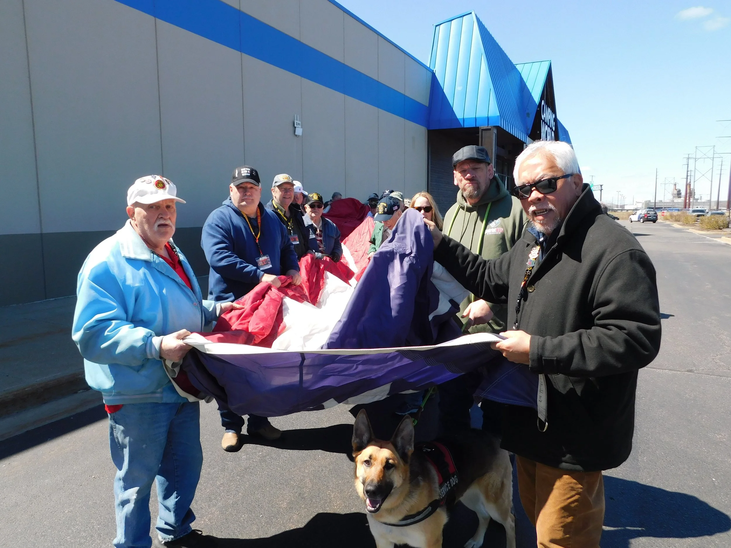 Group of people standing outside holding a large flag or banner, with a dog on a leash in the foreground, in front of a building with a blue roof and a parking lot.