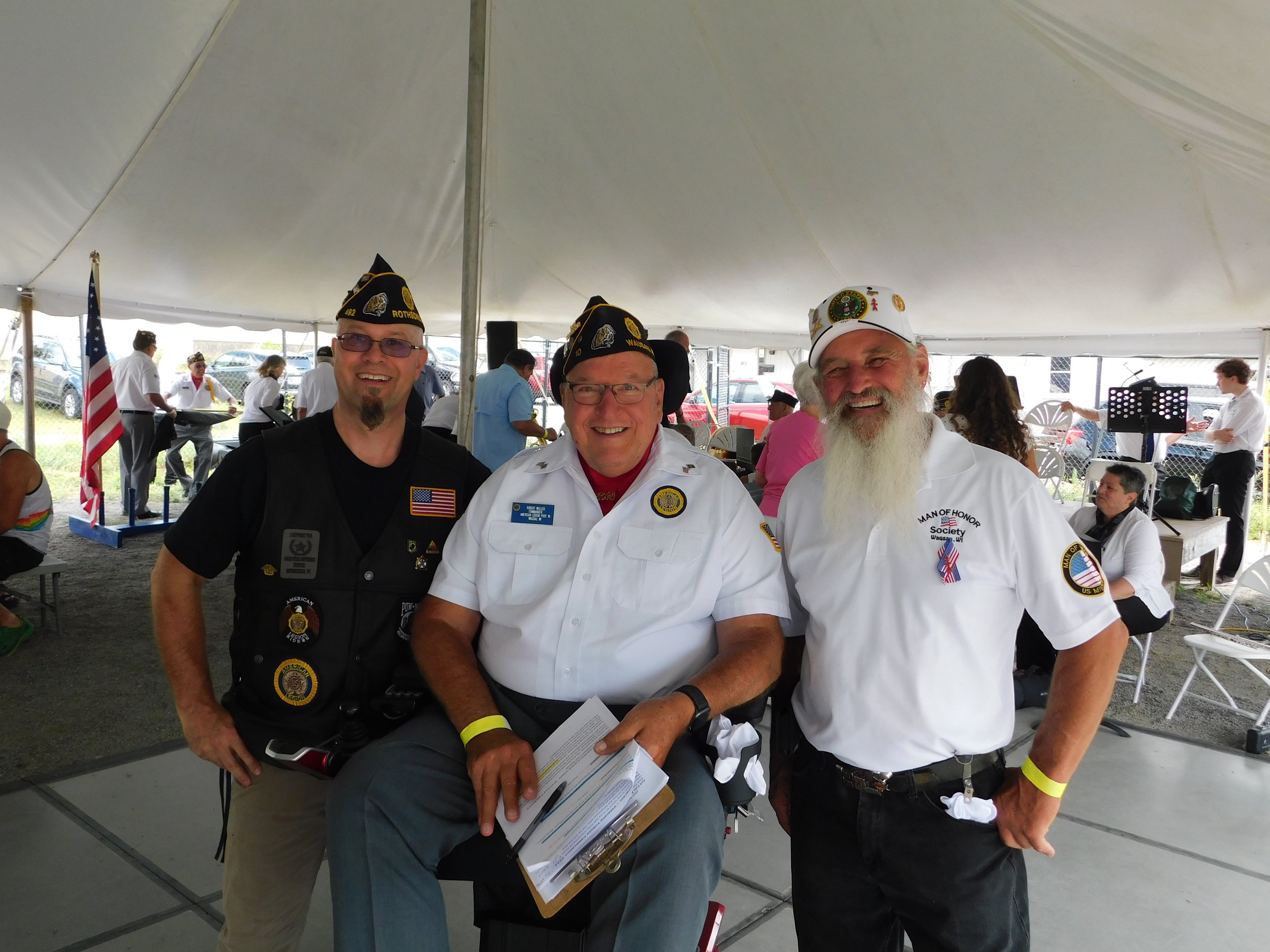 Three men dressed in VA and VFW uniforms standing under a large white tent, smiling at the camera, during an event.