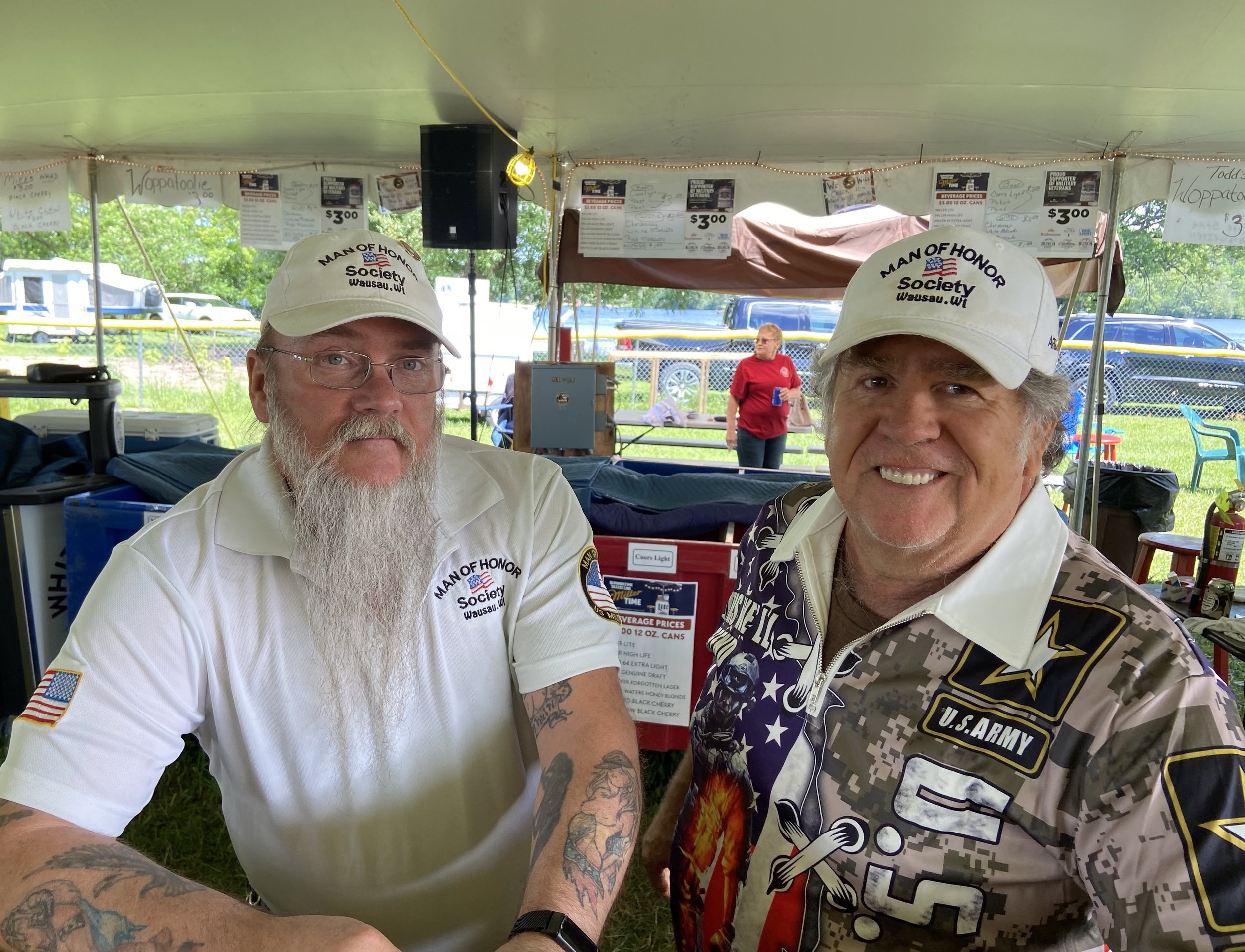 Two men wearing white 'Man of Honor Society' hats and shirts, one with a long beard and tattoos, smiling at the camera under a tent at an outdoor event.