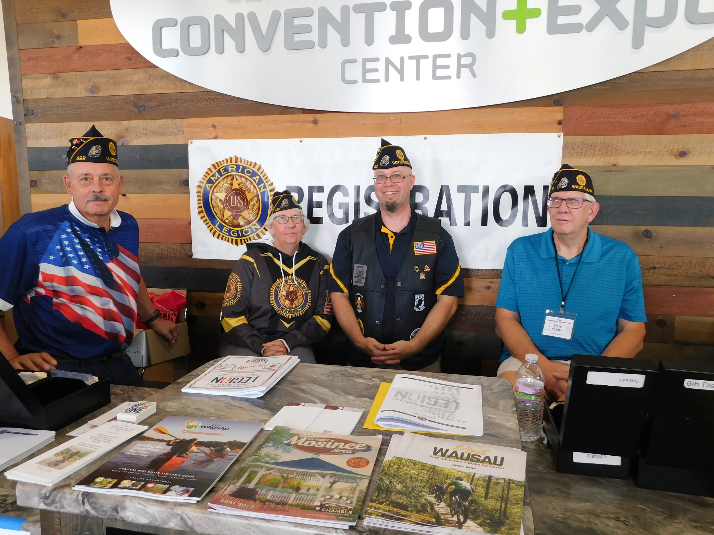 Four people wearing American Legion hats standing behind a registration table at a convention center. The table has brochures and papers. A banner with the American Legion logo and another with 'Registration' are behind them.