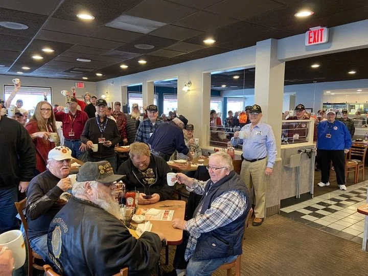 Veterans gathered in a restaurant, some wearing military caps, sitting at a table and socializing, while others are standing and posing for the photo.