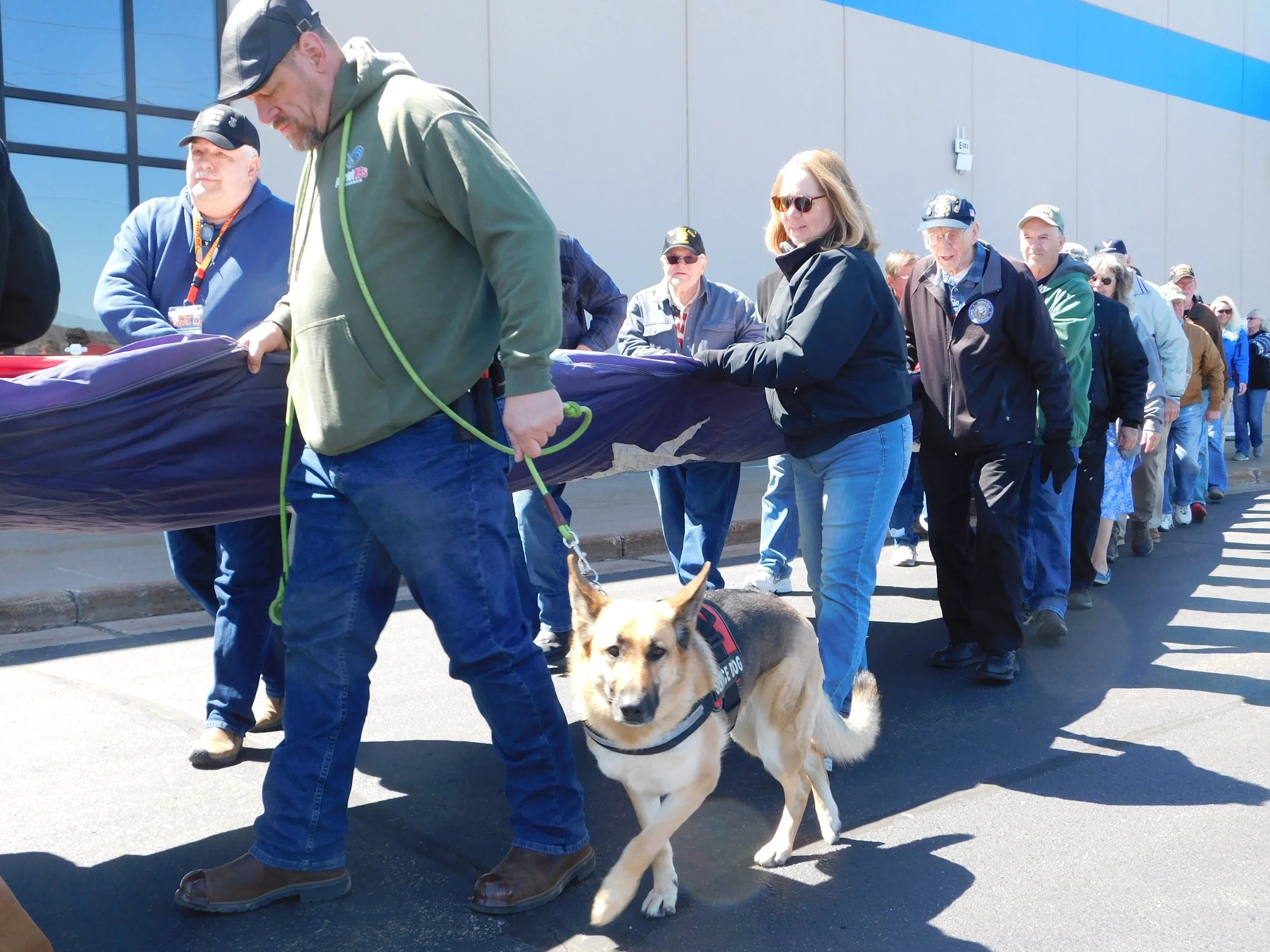 A group of people in a line participating in a parade or event outside a building, some are wearing jackets and hats, a man in the front is holding a large dog on a leash, and a woman nearby is helping hold a long object covered with a cloth.