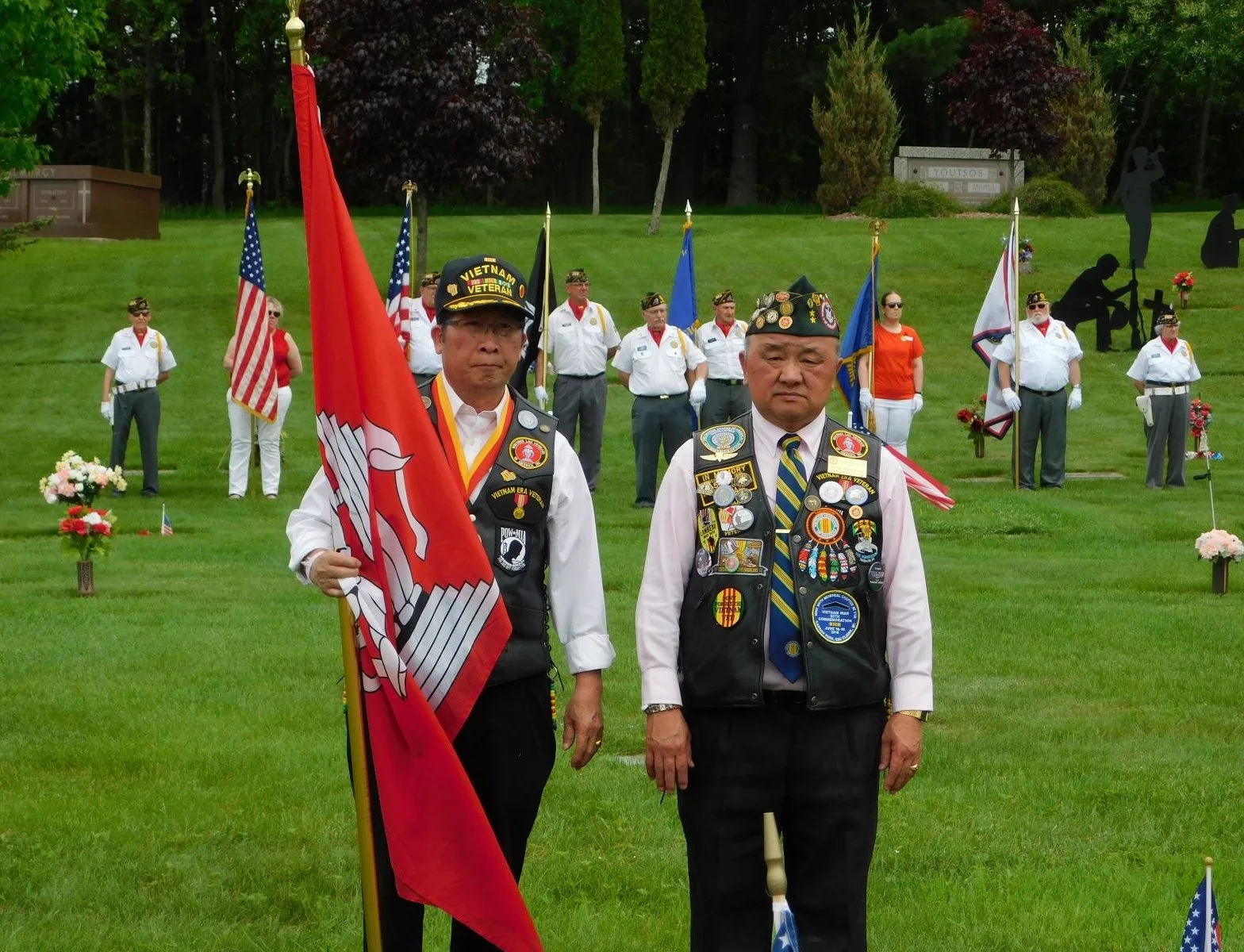 Members of a veterans' memorial service holding flags, with two in the foreground wearing veteran vests decorated with medals. One holds a red flag, and the other has a cap with military insignia. Several other veterans stand behind them, some holdin
