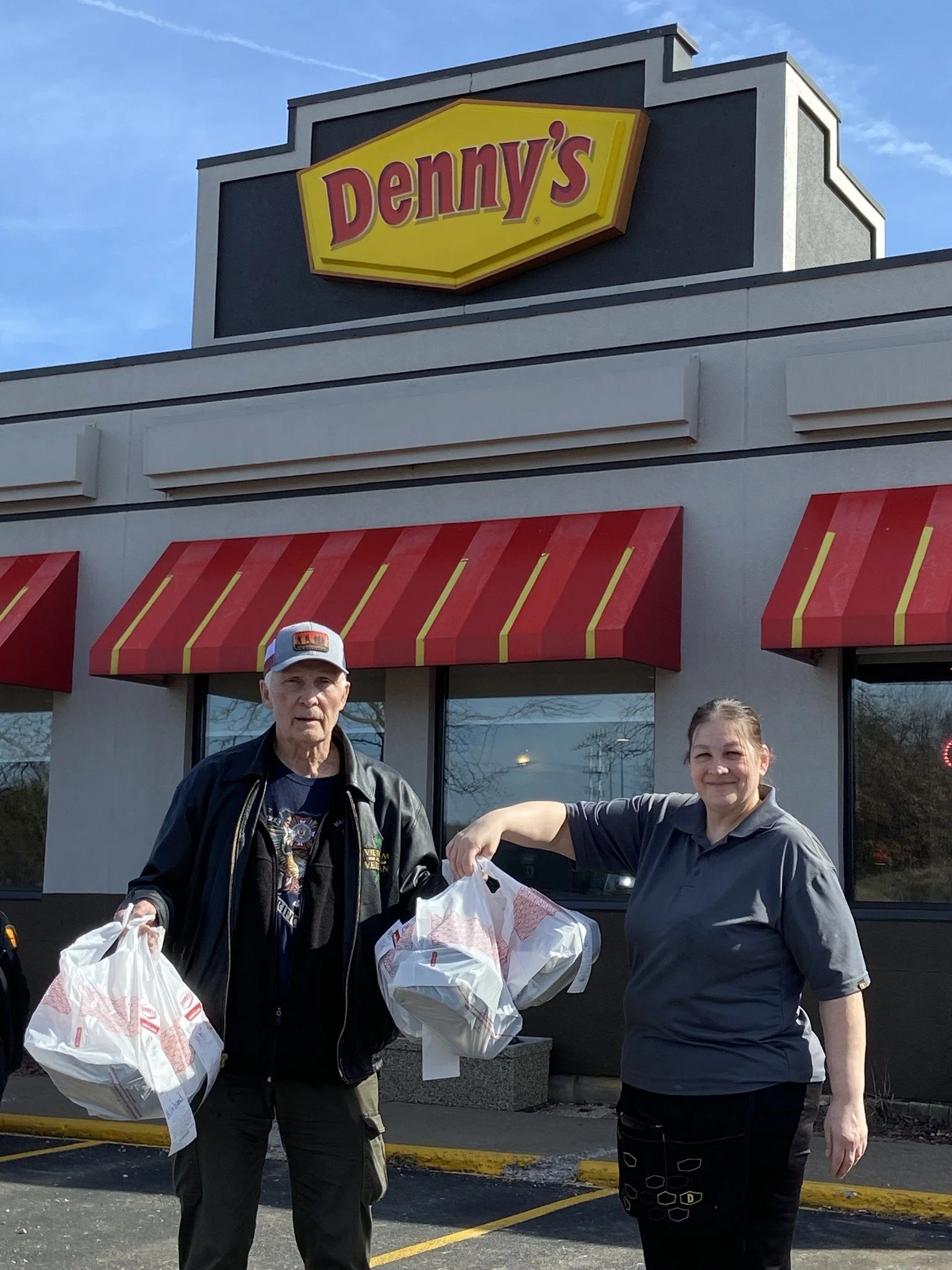 Two people standing outside a Denny's restaurant, each holding plastic bags of food, with a clear blue sky in the background.
