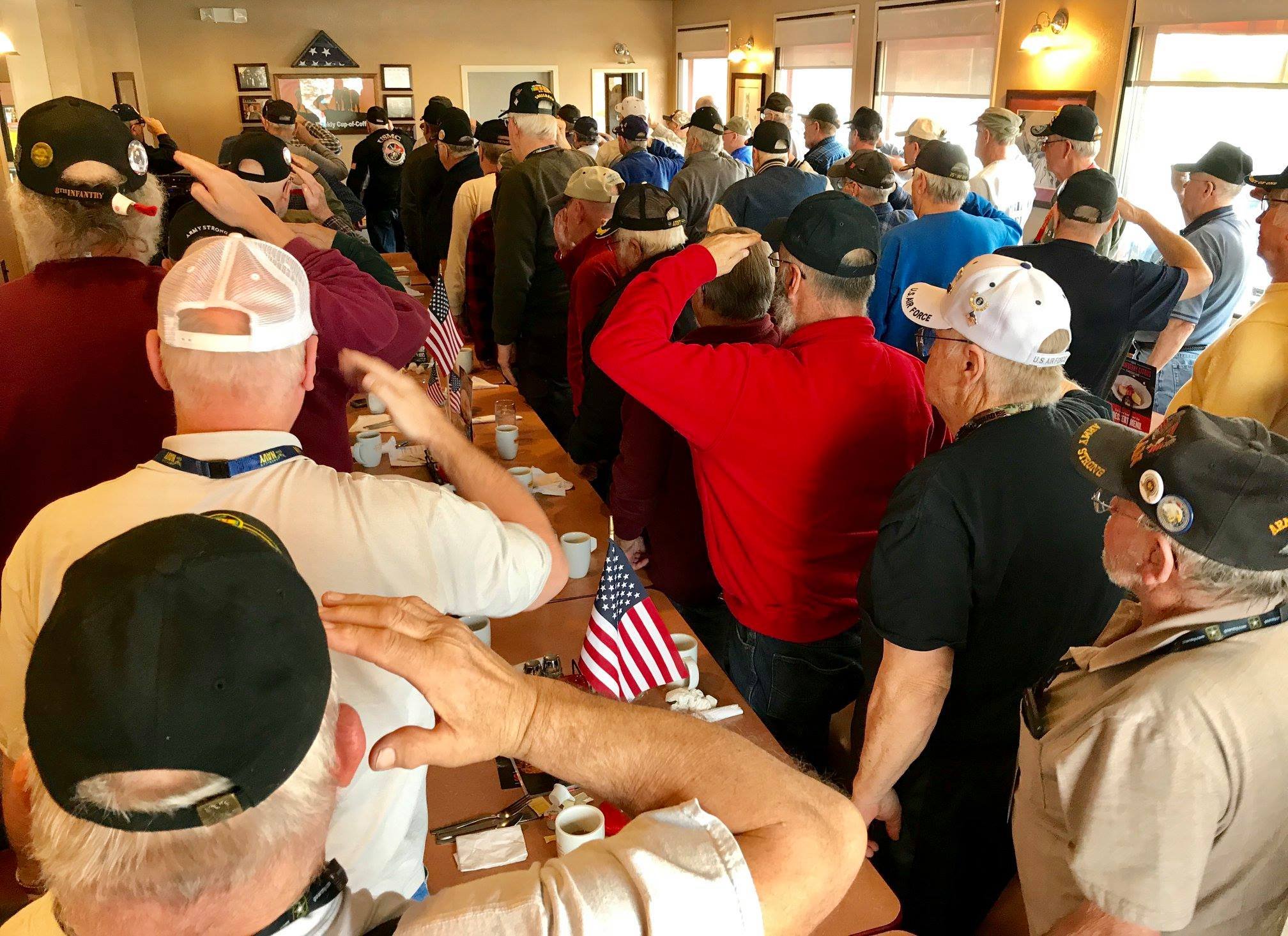 A group of military veterans, mostly older men, standing together in a room, some saluting or saluting, with American flags on the table and a patriotic atmosphere.