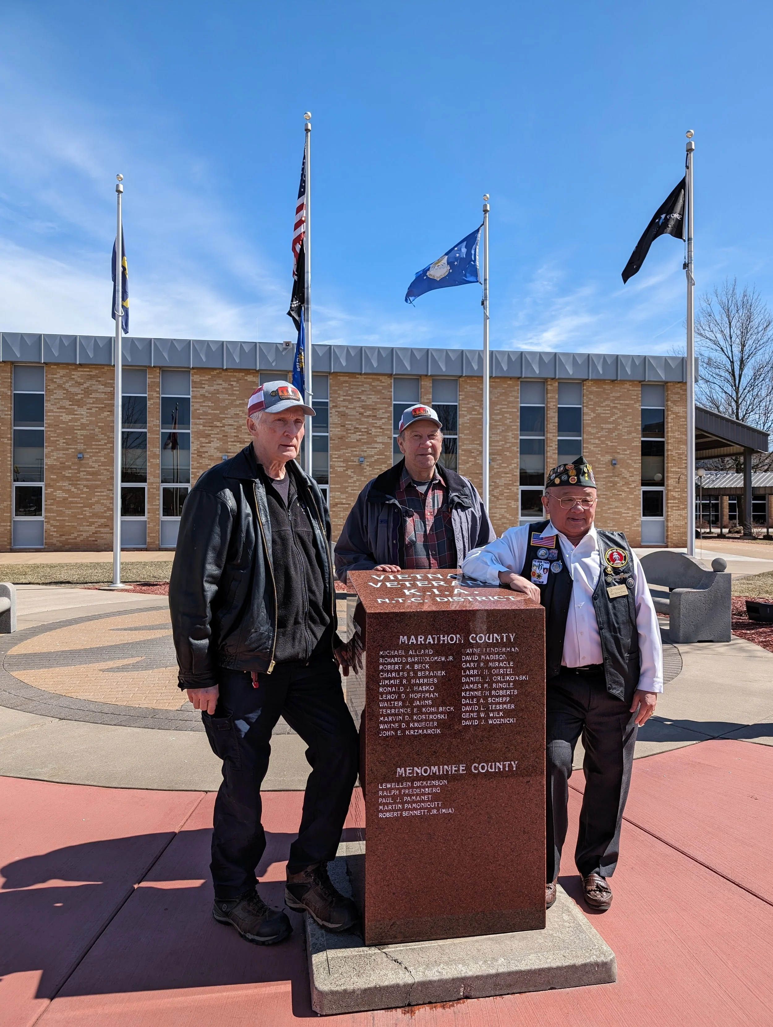 Three men standing beside a memorial plaque at a Veterans and Memorial Day event, with flags flying behind them, in front of a brick building on a sunny day.