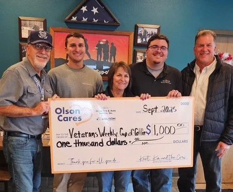 Five people standing together holding a large check for $1,000 made out to Veterans Weekly Cup of Coffee, dated September 2023, in an indoor setting with patriotic decorations and framed photos in the background.