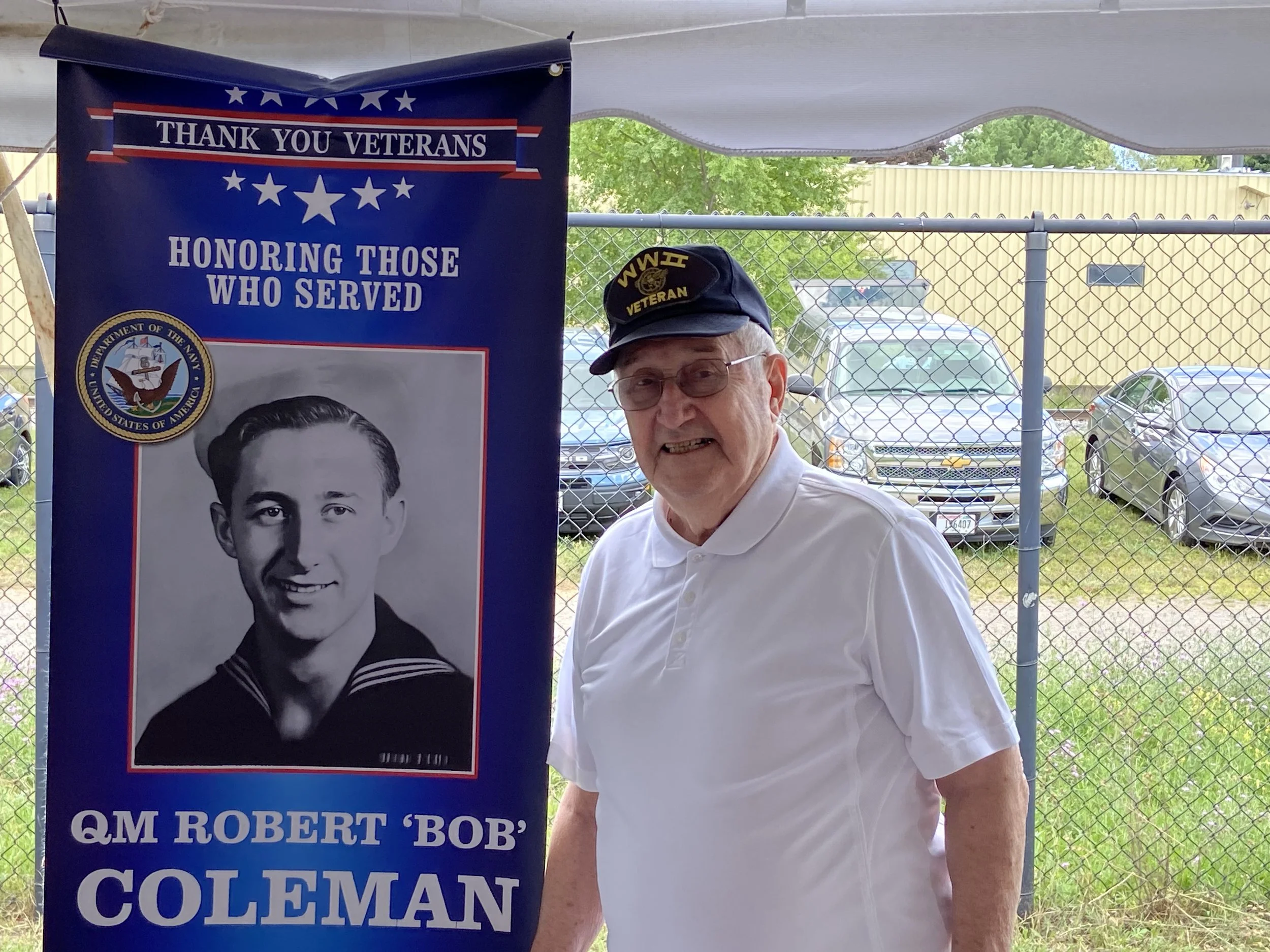 An elderly man wearing a navy veteran cap and glasses, standing beside a large blue banner that honors veterans. The banner features a black and white portrait of a young man in a Navy uniform, with the text: 'THANK YOU VETERANS, HONORING THOSE WHO S
