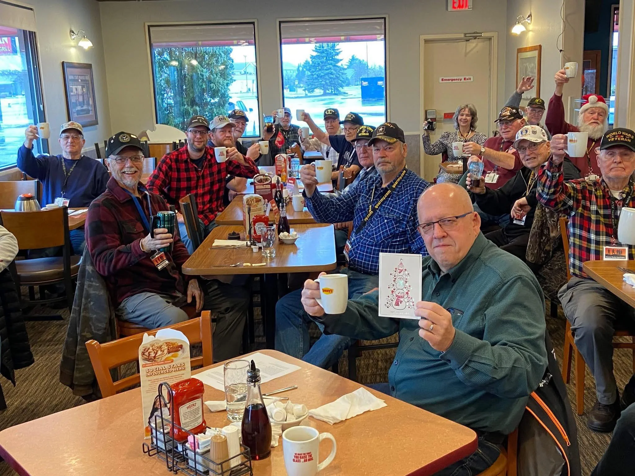 Group of people, mostly veterans wearing caps, sitting at tables in a restaurant, raising mugs in a toast, celebrating together.