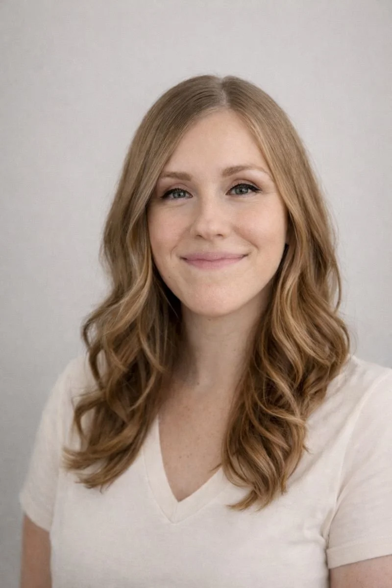 A woman with light brown, wavy hair wearing a white V-neck t-shirt, smiling at the camera against a plain, light-colored background.