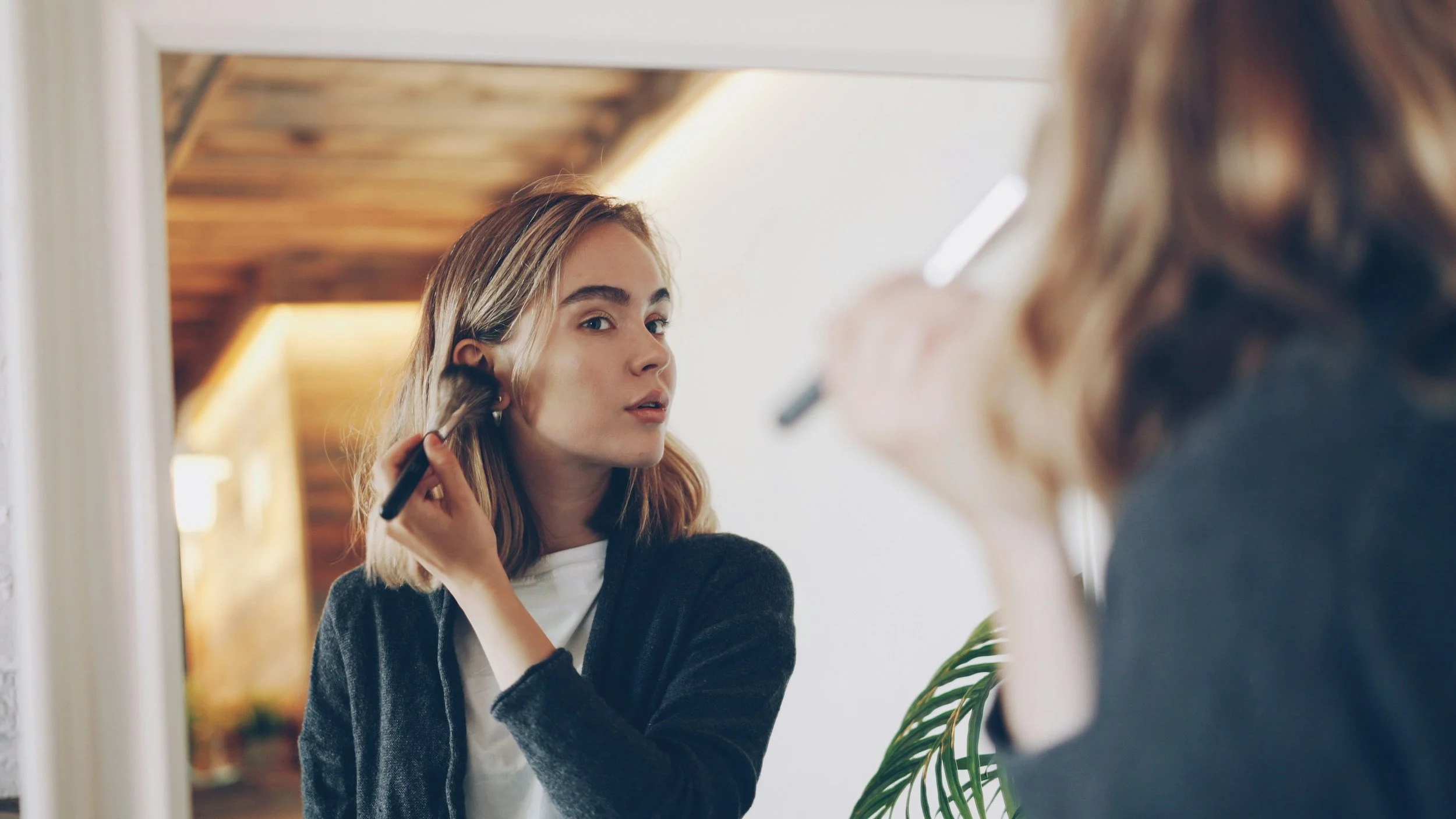 A woman with shoulder-length blonde hair applies makeup using a brush while looking into a mirror.