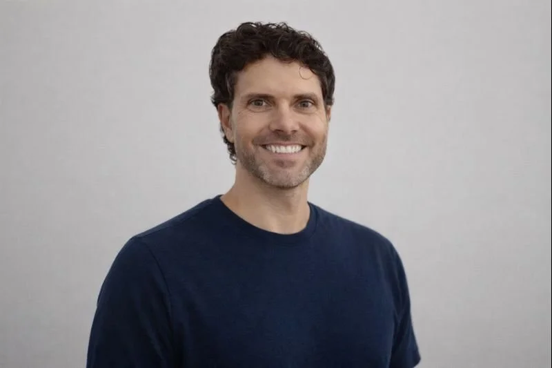 Smiling man with curly brown hair and beard, wearing a navy blue shirt, standing against a light gray background.