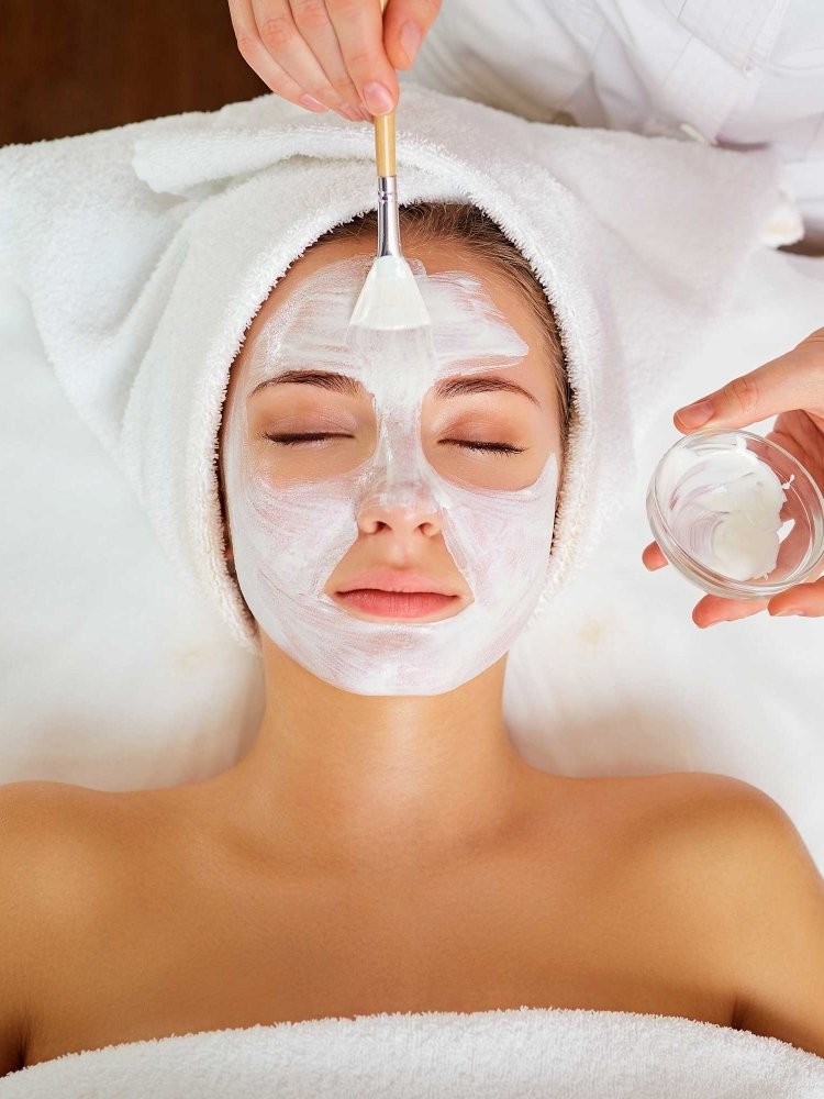 Woman receiving a facial treatment while lying down with eyes closed, a white towel wrapped around her head, and a white facial mask being applied by a skincare professional.