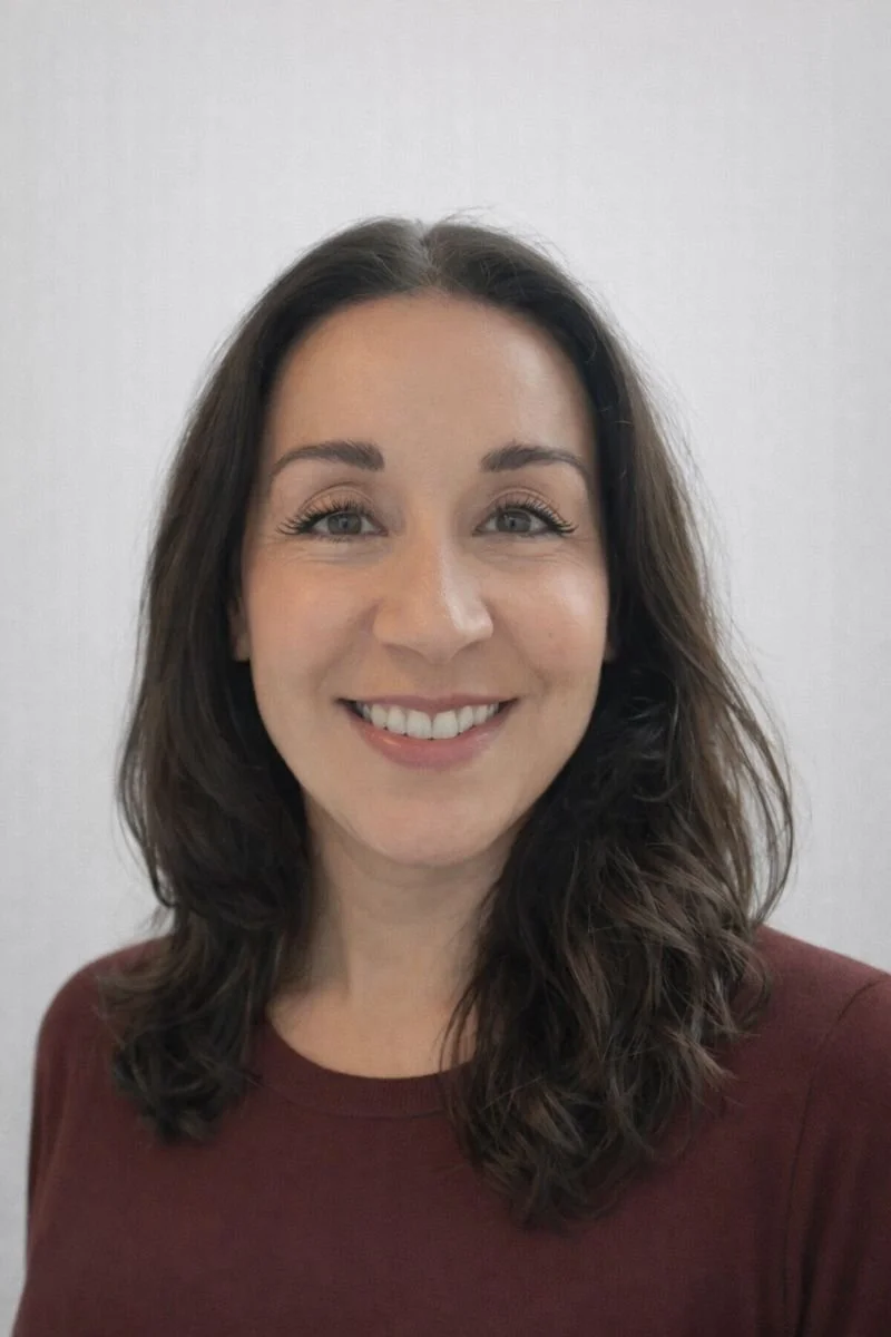 A woman with shoulder-length wavy brown hair, light skin, wearing a maroon top, smiling against a plain light background.