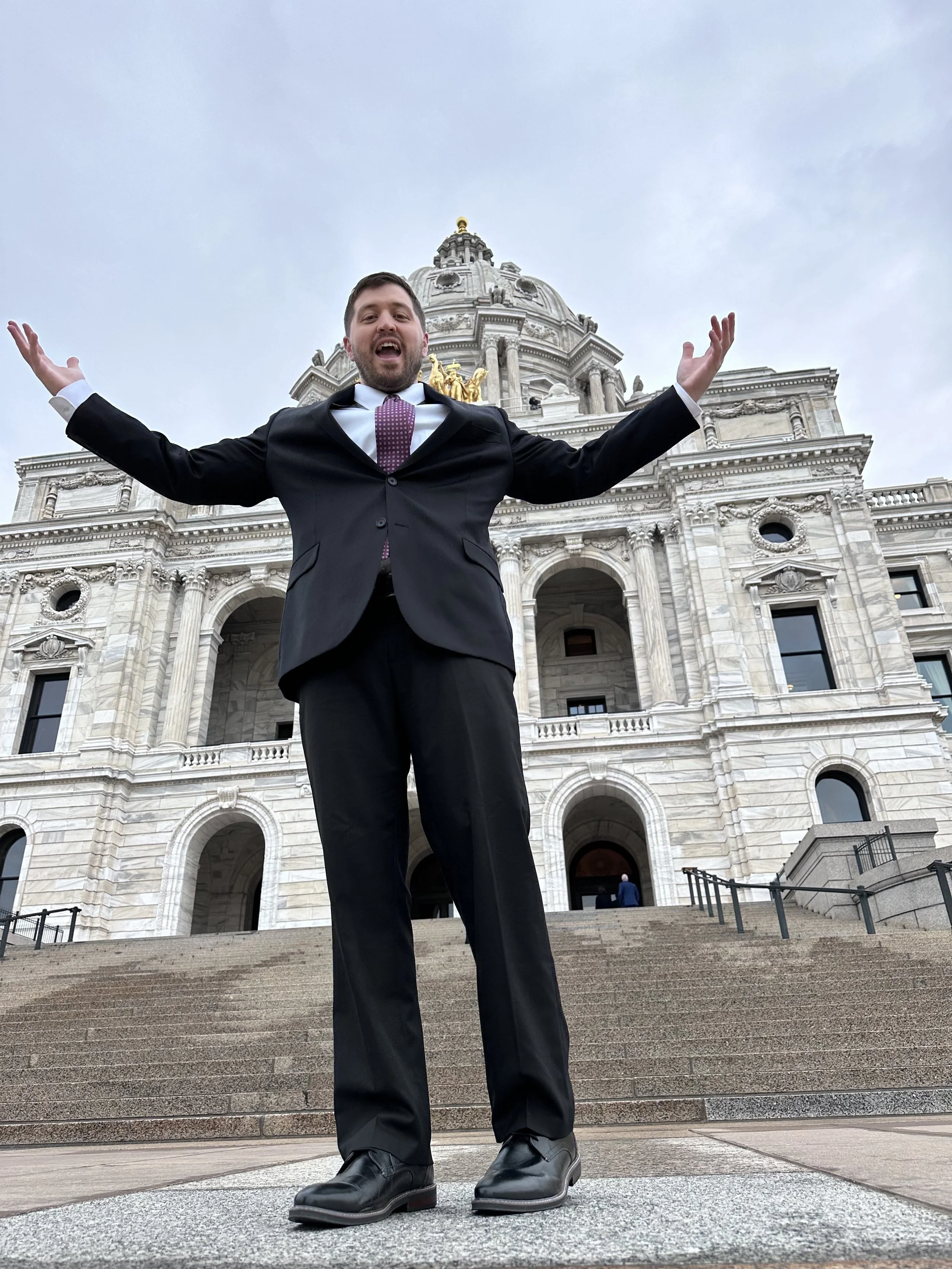 A man in a black suit and purple tie standing on steps in front of a large, ornate government building, with arms outstretched and smiling.