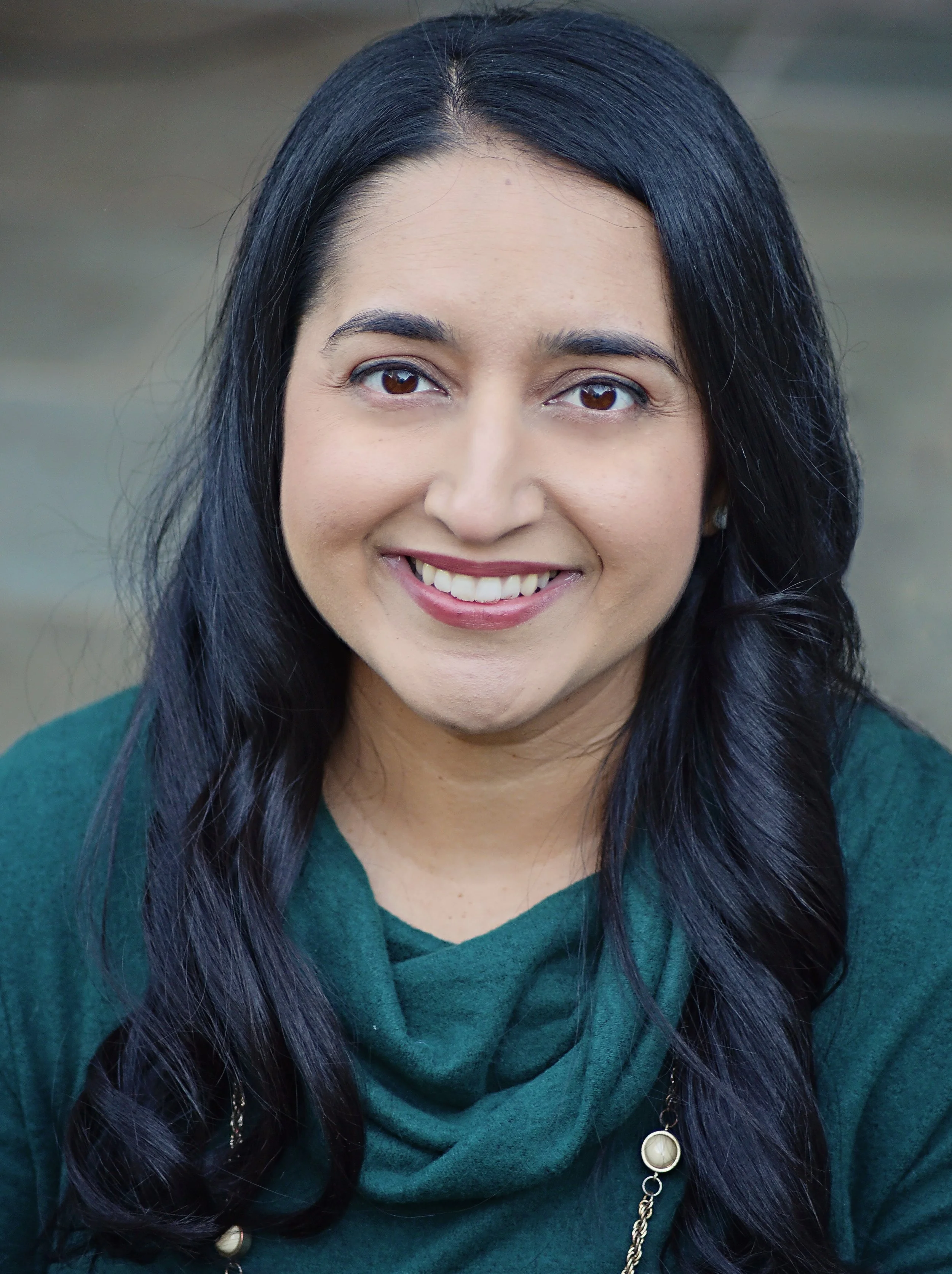 Headshot of a smiling woman with long dark hair, wearing a green top and necklace, outdoors with blurred background.